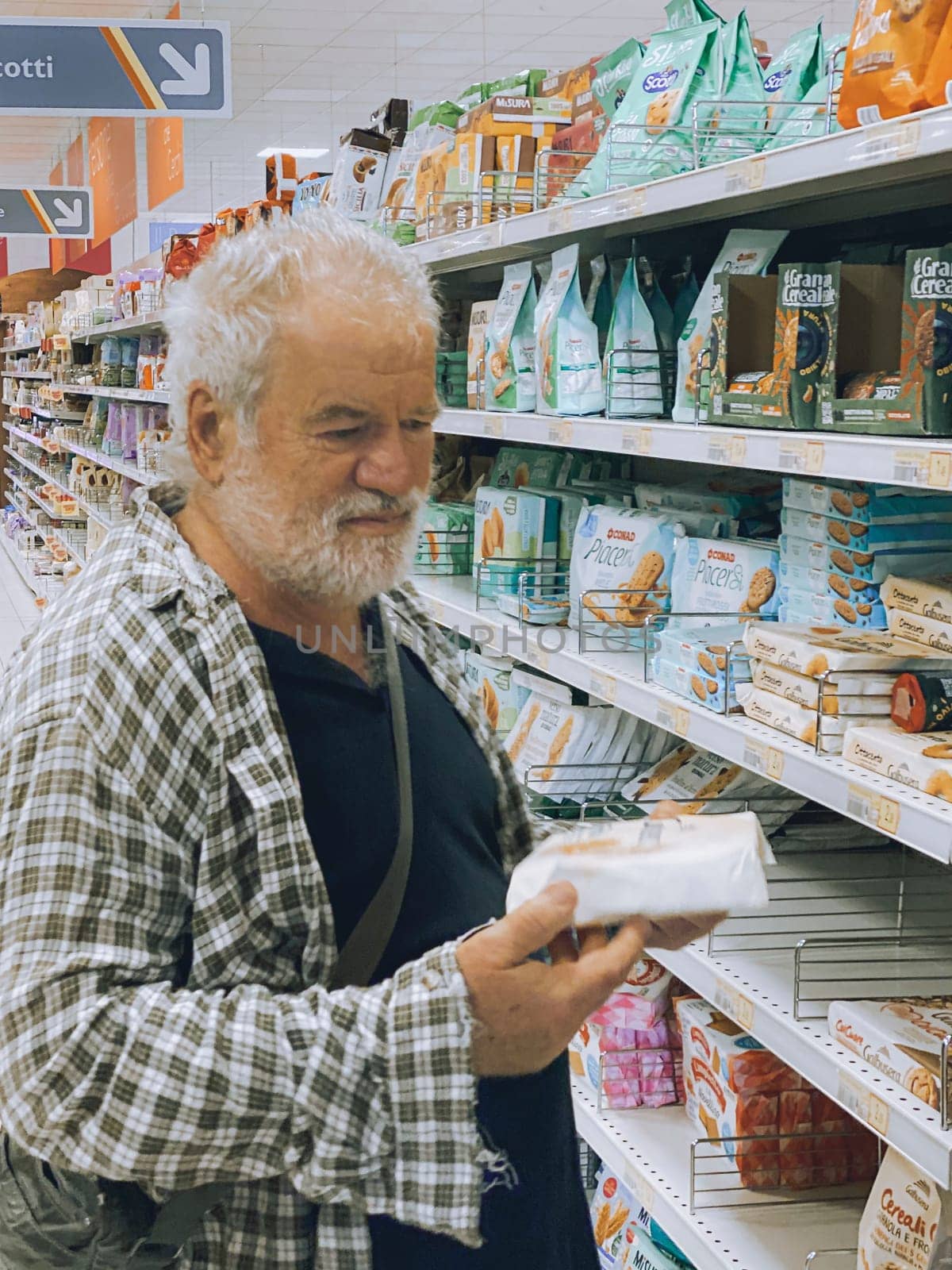 Cremona, Lombardy, Italy October 15th 2024 Elderly man shopping for cookies in a supermarket, carefully checking the packaging information before making a purchase