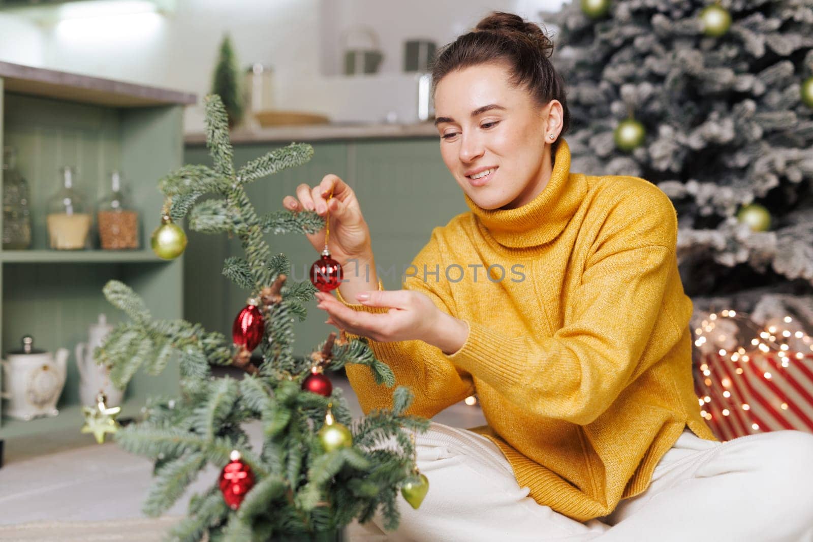 Woman in yellow sweater decorating small Christmas tree with toys in cozy home atmosphere. merry christmas