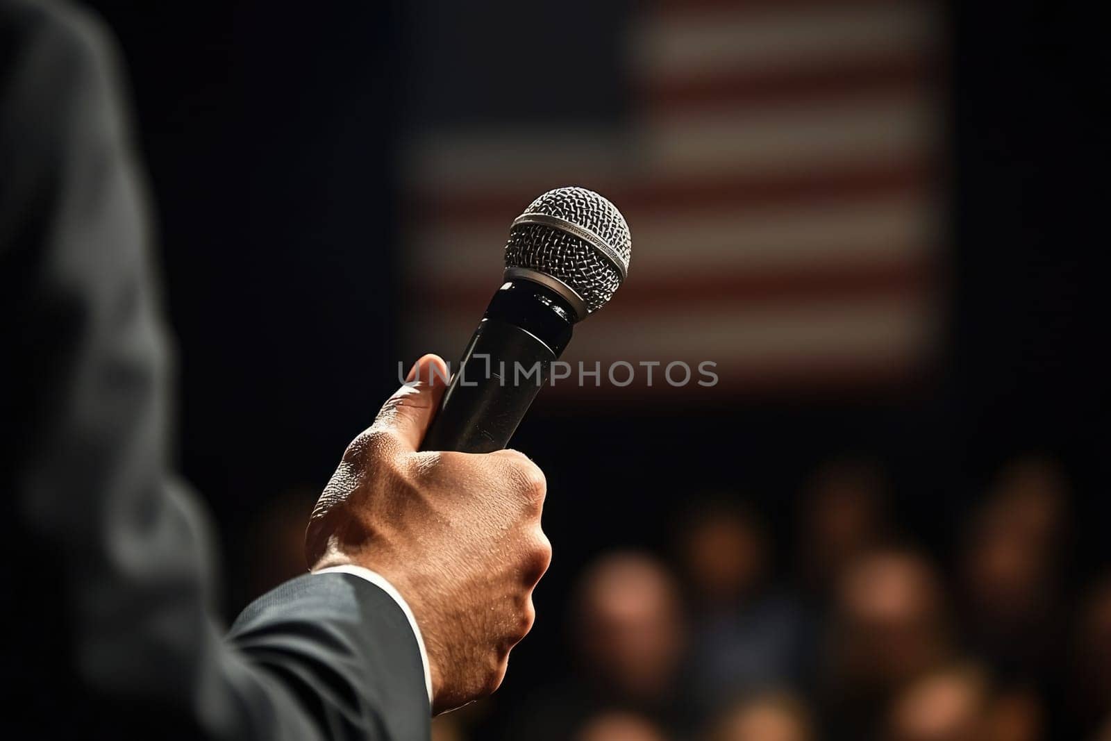 A close-up image of a professional speaker's hand holding a microphone, with an audience in the background and an American flag, symbolizing impactful public speaking.