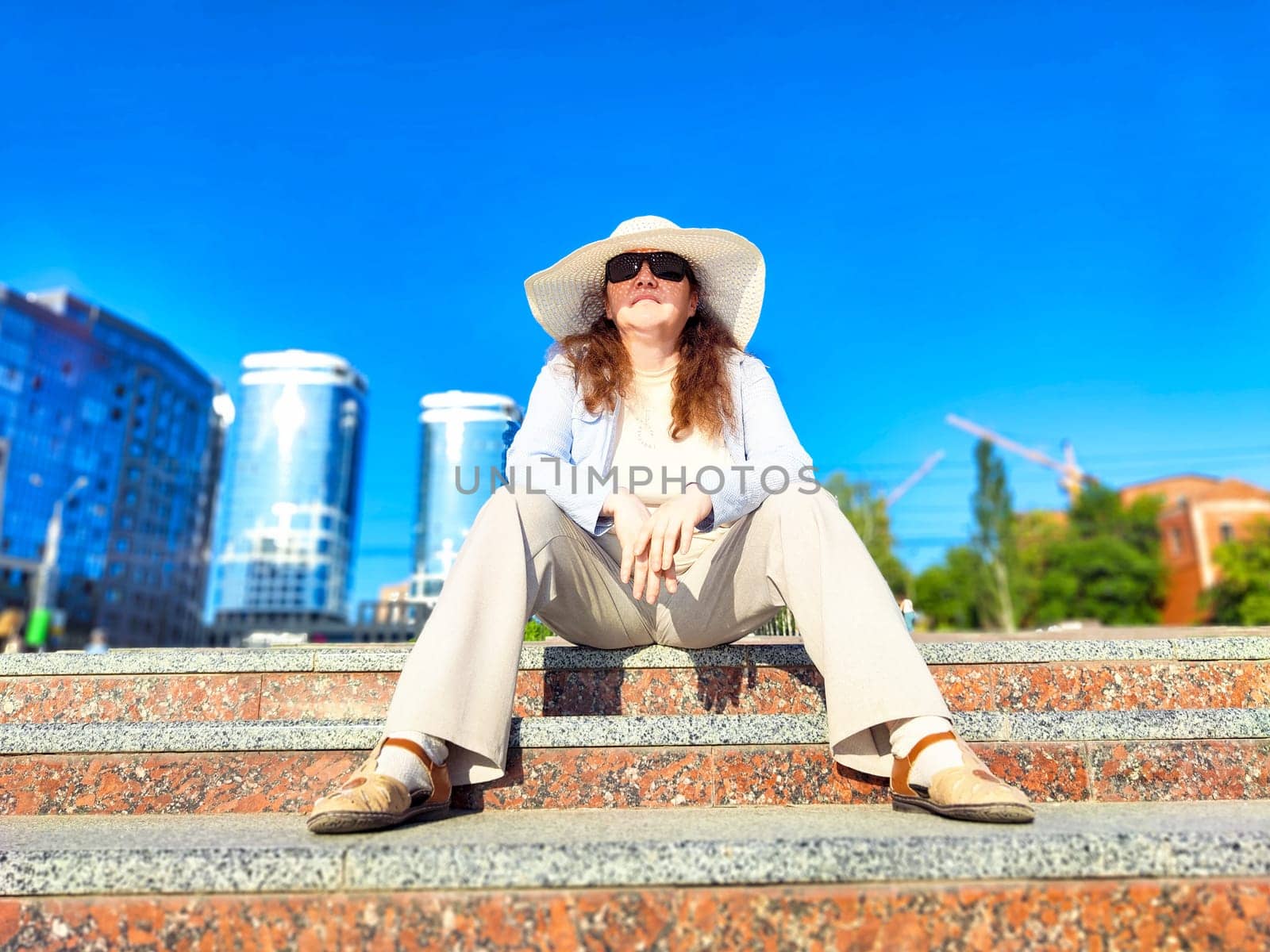 A woman sits comfortably on stone stairs in city, enjoying the warmth of a sunny day while wearing a wide-brimmed hat