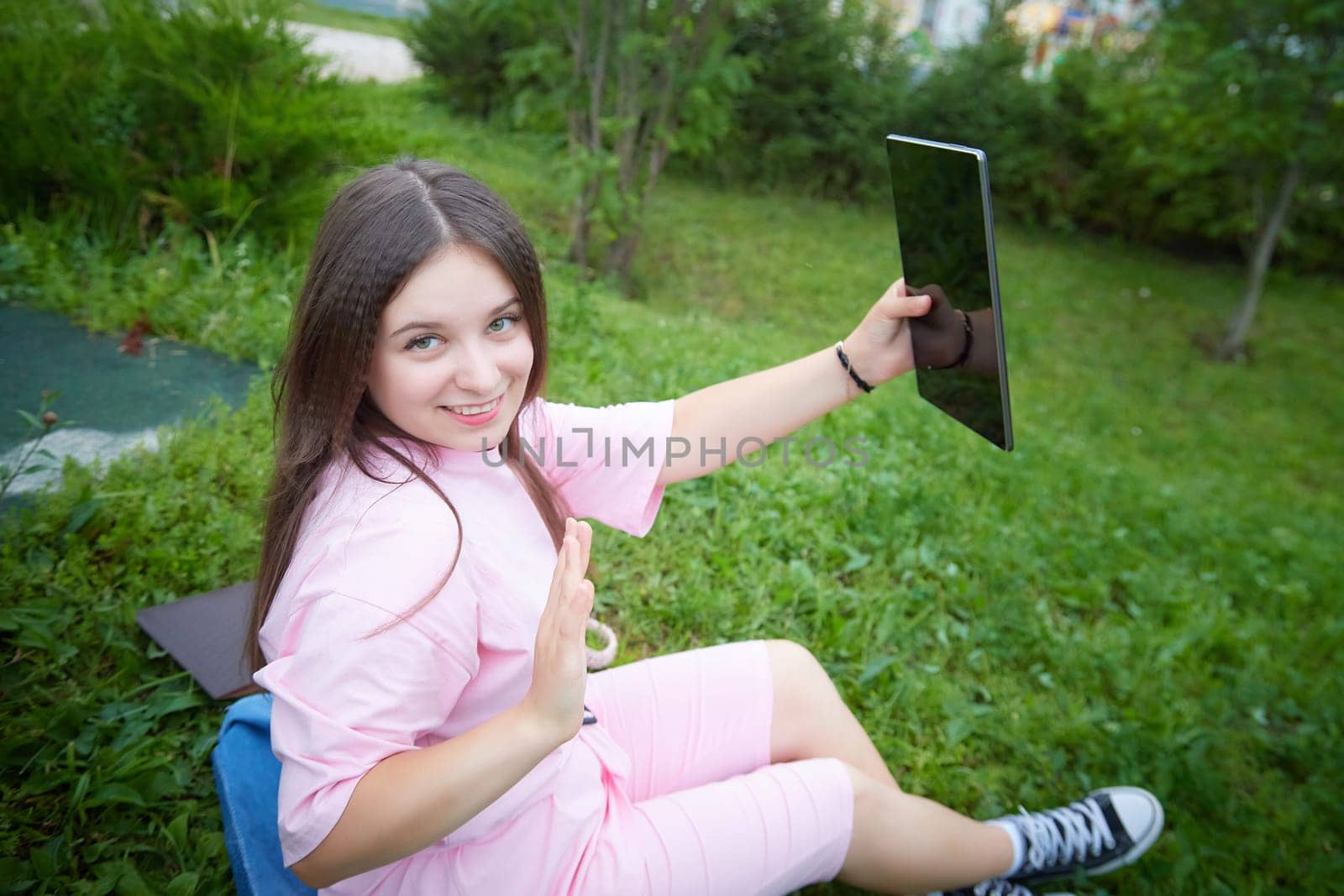 woman smiles as she looks at her tablet, sitting on grassy view and taking selfie. Blogger make podcast