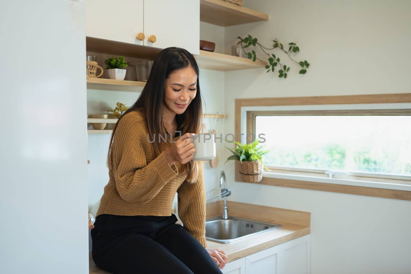 Smiling pretty woman enjoying a cup of coffee while sitting on kitchen counter.
