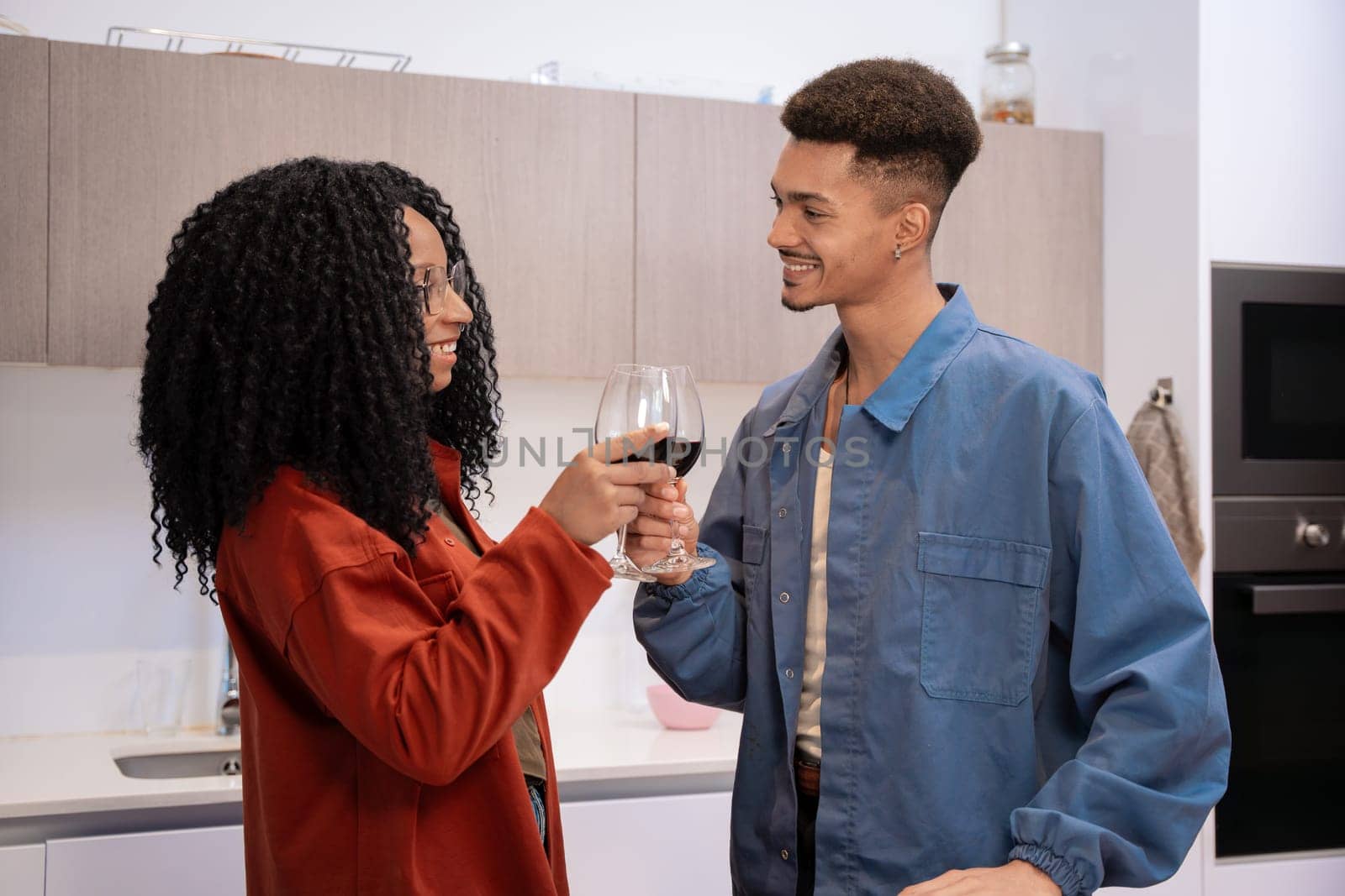 A young, stylish couple smiling and toasting with glasses of red wine in a modern kitchen. They wear casual, colorful clothing, creating a warm, joyful atmosphere. Perfect for lifestyle concepts