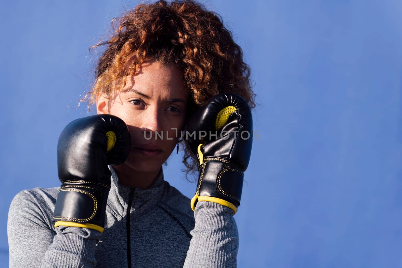 determined adult female boxer with curly hair ready to fight on boxing gloves during workout on blue background, concept of strength and healthy lifestyle, copyspace for text