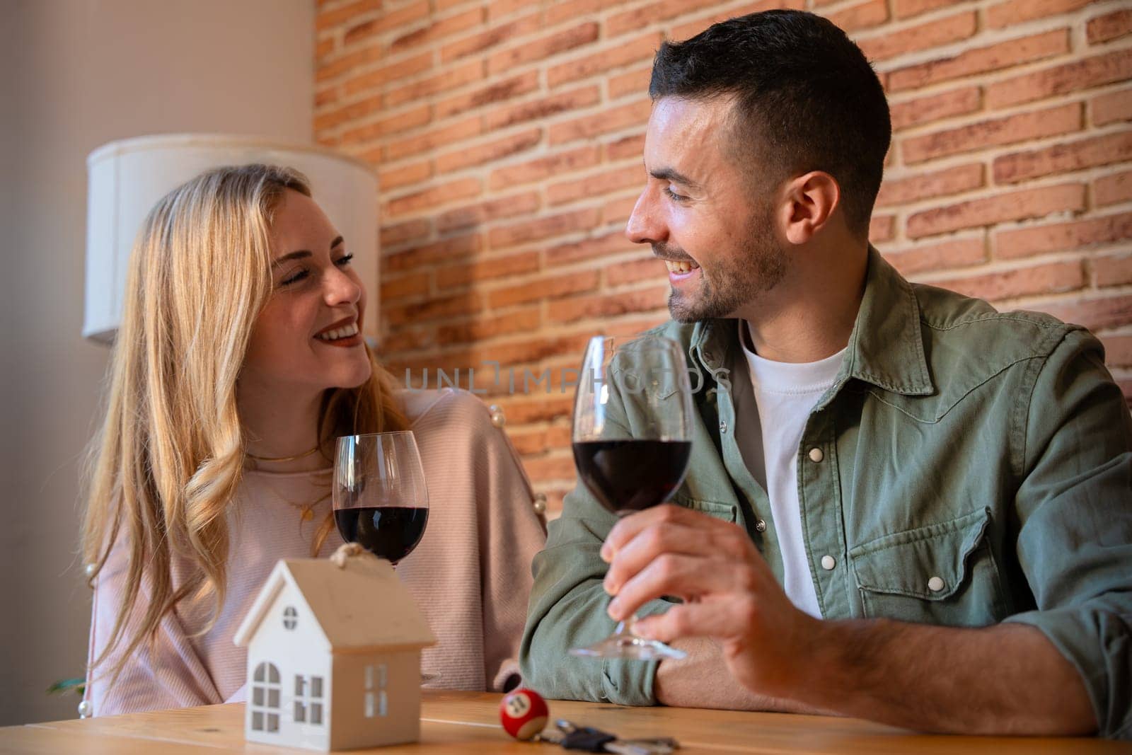 A young, stylish couple smiling and toasting with glasses of red wine in a modern kitchen. They wear casual, colorful clothing, creating a warm, joyful atmosphere. Perfect for lifestyle concepts