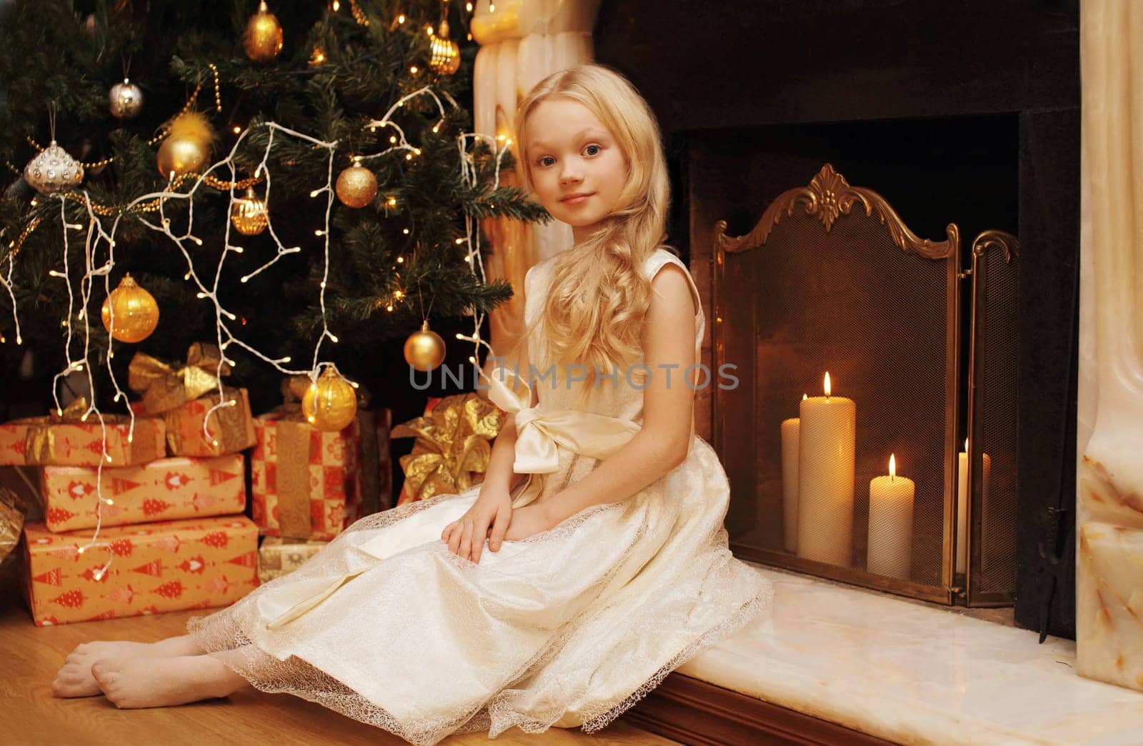 Happy little girl child in dress with festive gift box sitting near Christmas tree and fireplace with holiday lights at home