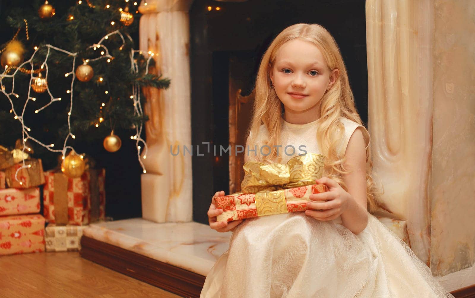 Happy little girl child in dress with festive gift box sitting near Christmas tree and fireplace with holiday lights at home