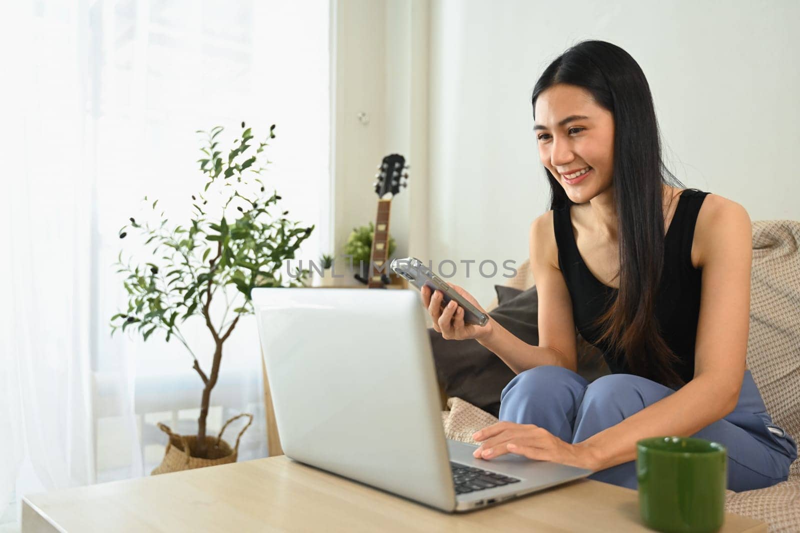 Cheerful young woman working from home with laptop and using mobile phone.