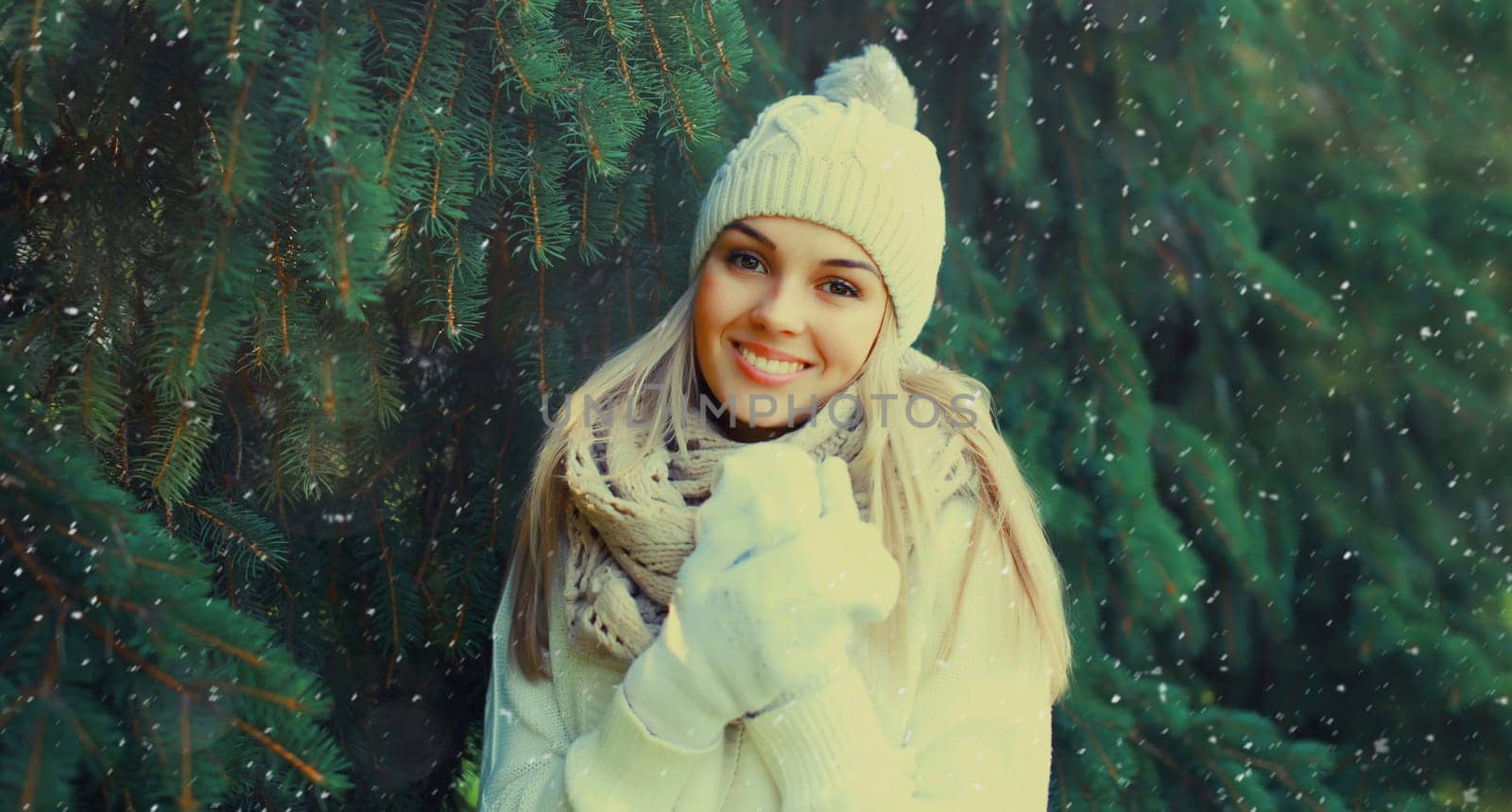 Winter portrait of happy smiling blonde young woman in forest with snow on Christmas tree background, wearing hat, enjoying warm weather, knitted sweater and scarf
