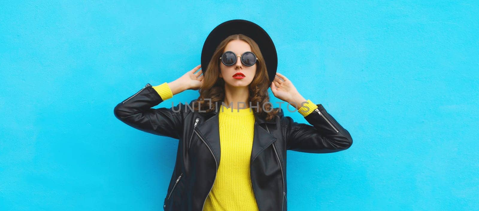 Fashion, trendy, stylish young woman in black round hat, leather jacket, yellow sweater, modern girl posing on blue studio background