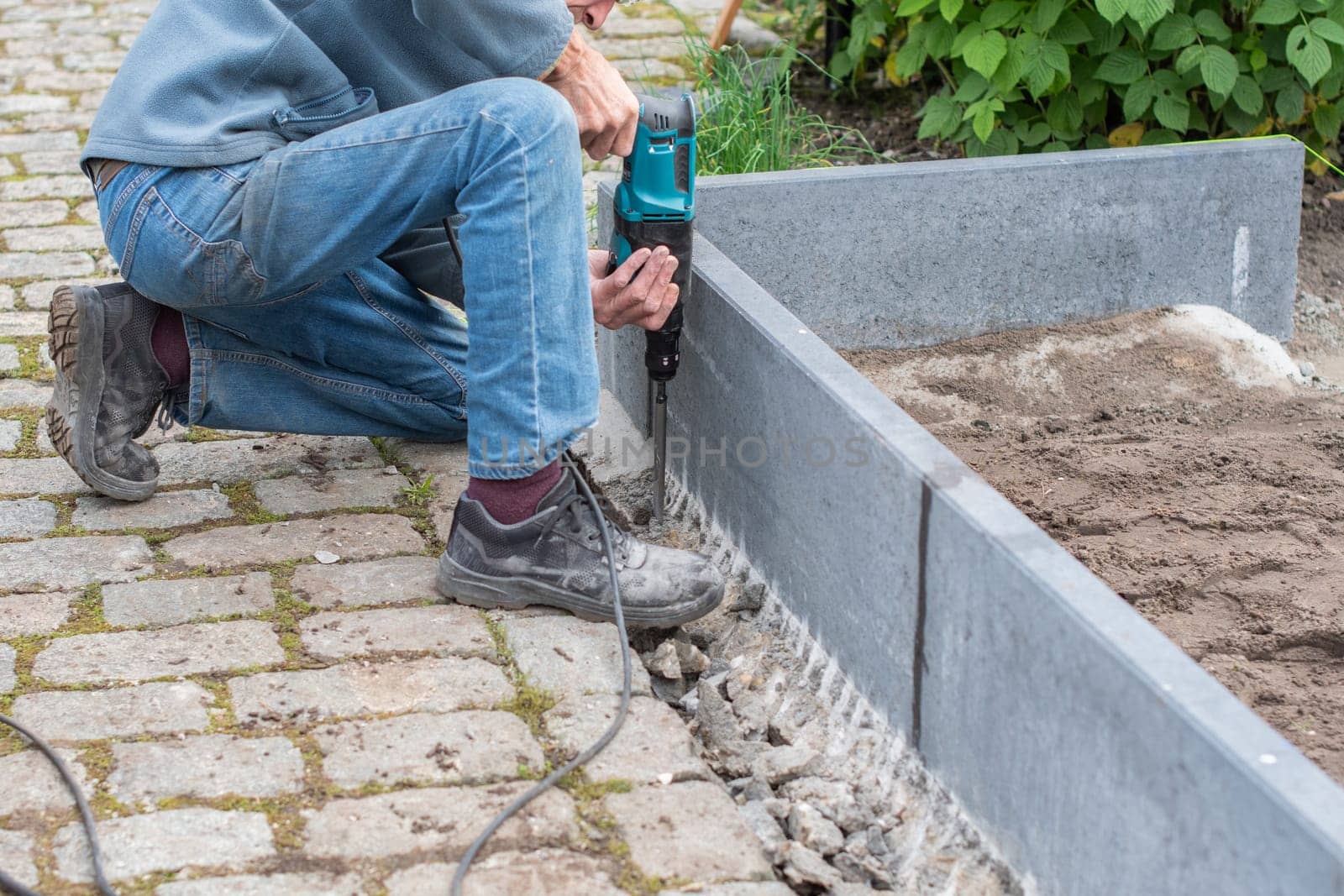 middle-aged man drills holes in the foundation of greenhouse for strengthening, improvement of garden or vegetable garden,frame for beds with soil,concept of home ecological turning of vegetables,