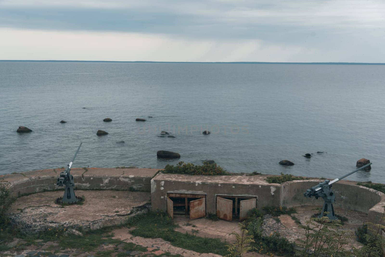 An abandoned coastal artillery bunker with cannons overlooks a calm sea under an overcast sky, evoking a sense of historical significance and quiet solitude in a rugged coastal landscape