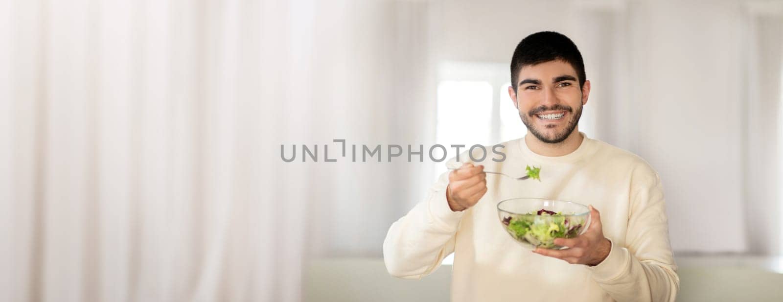 A smiling young man holds a bowl of fresh salad while taking a bite. The bright indoor space enhances the healthy lifestyle vibe, highlighting freshness and good nutrition.