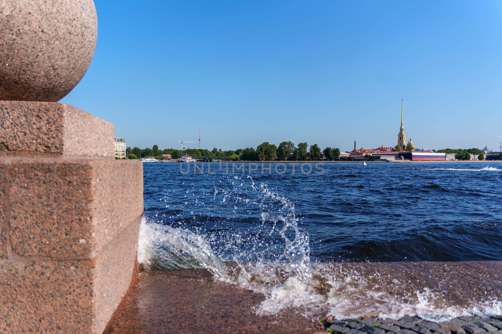 Close-up of waves crashing against a granite embankment on a sunny day, with a historic cityscape in the background under a clear blue sky. Vibrant waterfront scene