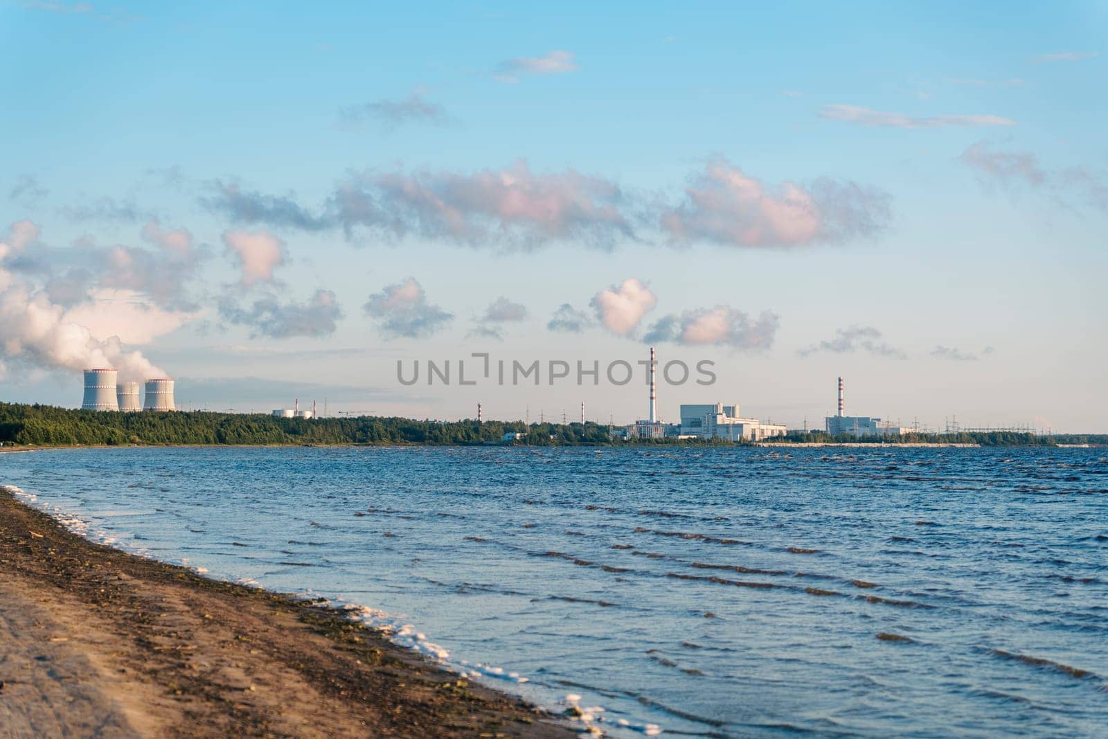 Industrial facility with smokestacks near a coastal shoreline, with blue sky and clouds, viewed from the beach. Waves gently reach the sandy shore in a calm, open scene
