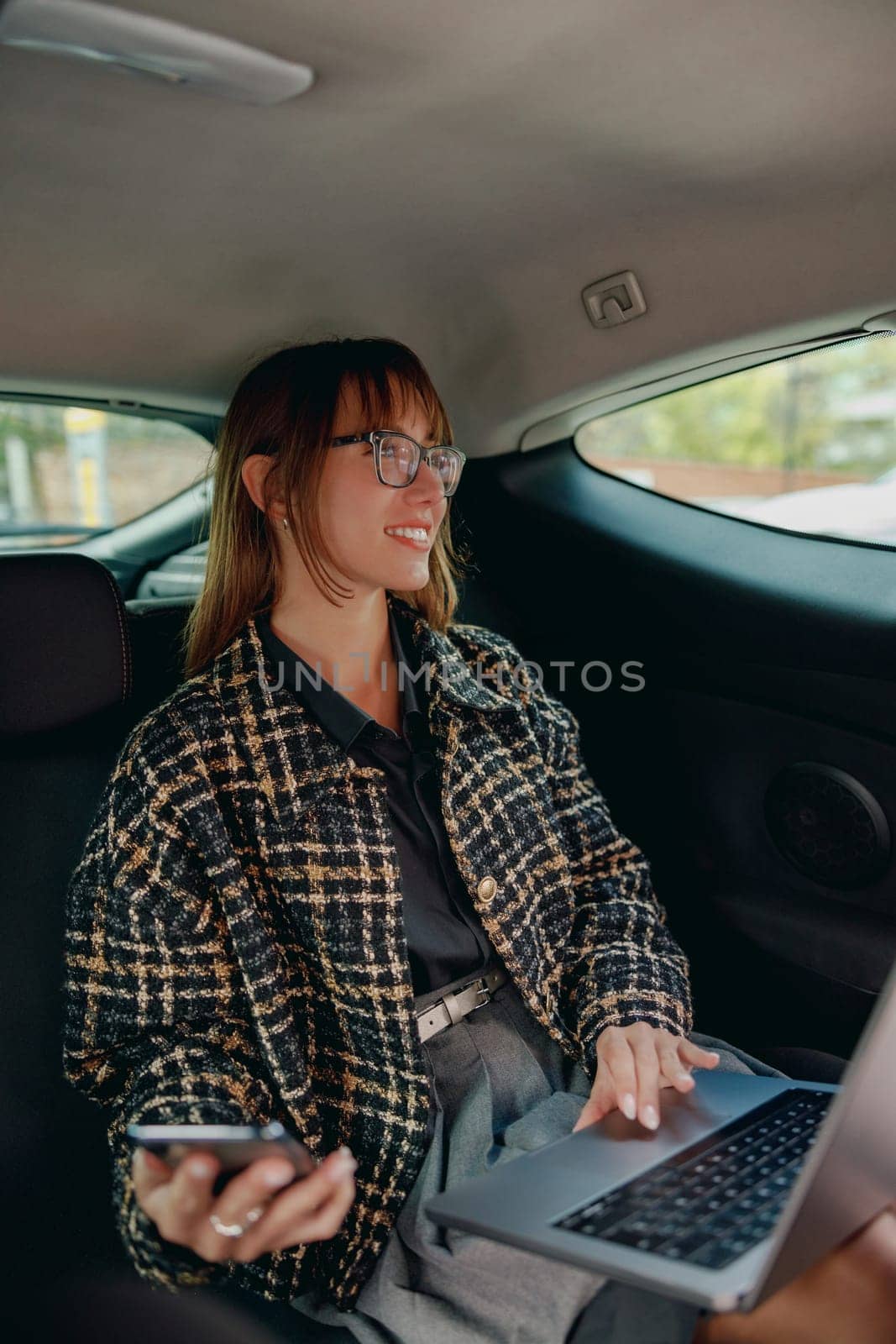 A young woman skillfully utilizes her laptop and smartphone inside a car, expertly showcasing her remote work lifestyle