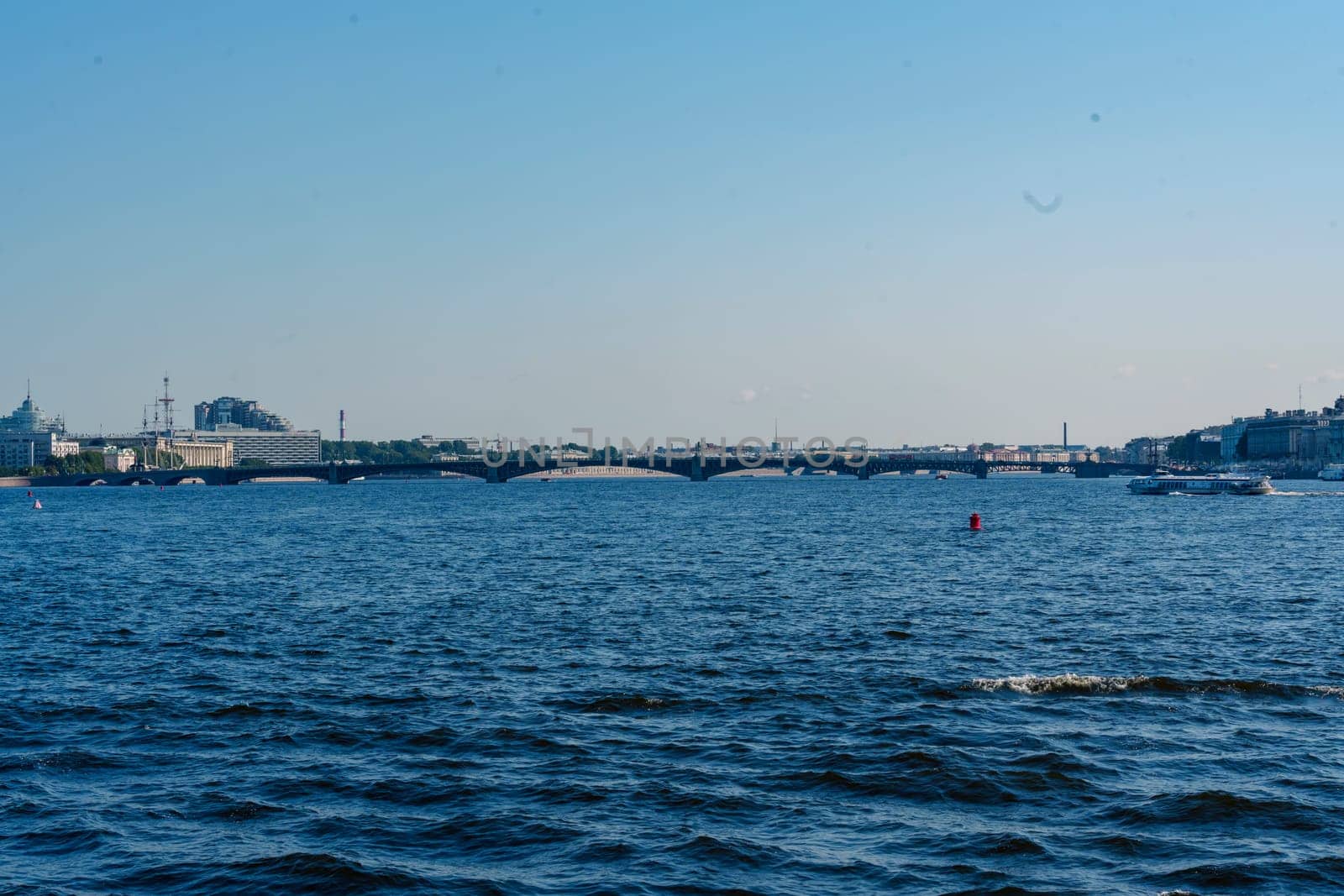 Scenic view of a wide river with a historic bridge spanning across, framed by urban buildings under a clear blue sky. Peaceful waterfront scene with gentle waves