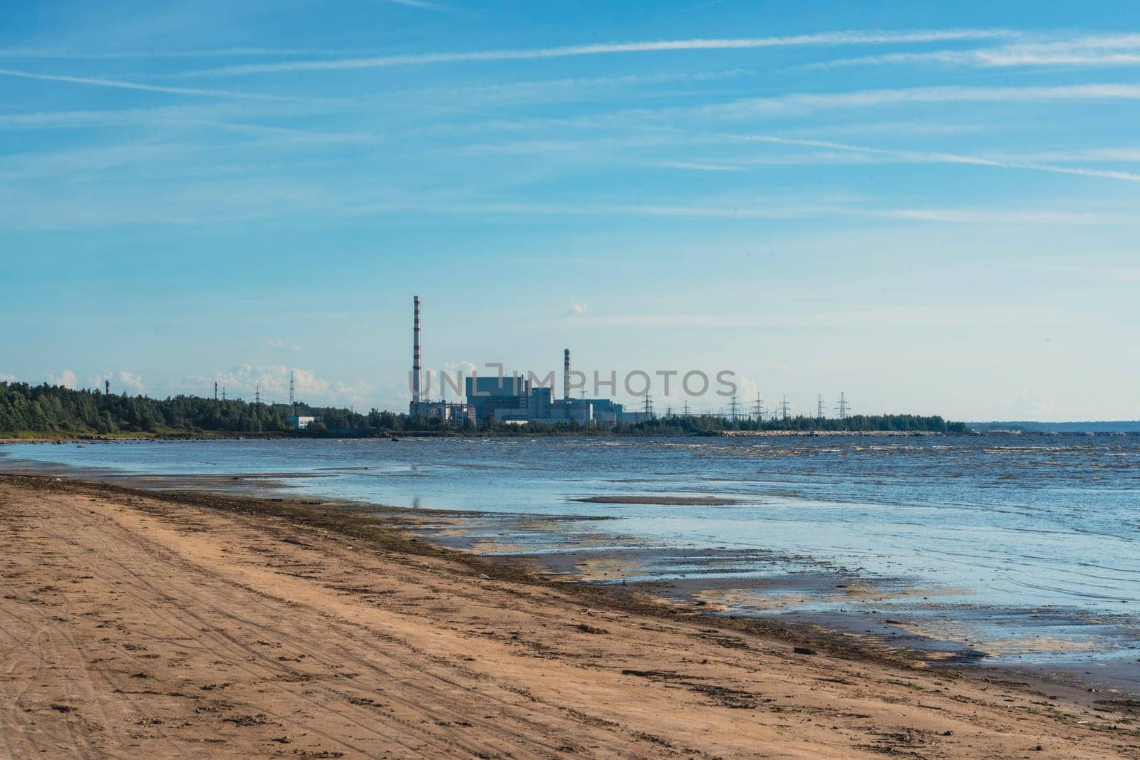 A power plant stands along a sandy beach under a clear blue sky, juxtaposing industrial structures with the natural coastal landscape on a sunny day