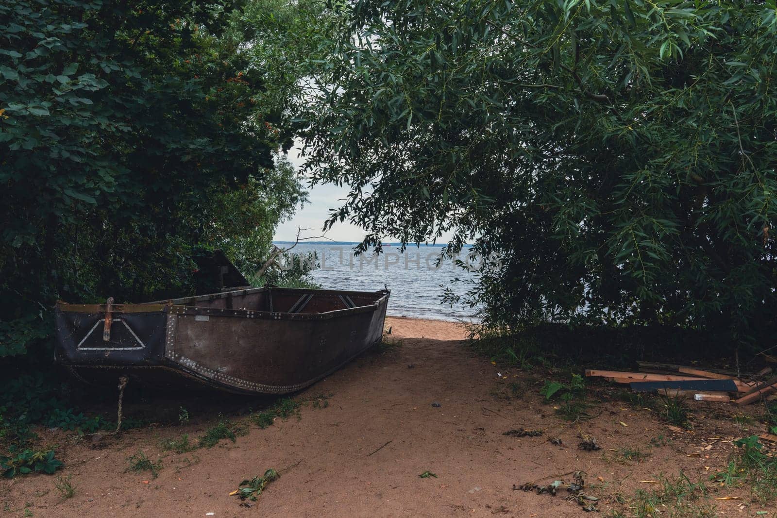 An old wooden boat rests on a sandy shore, partially hidden among trees, offering a secluded and peaceful setting by the water under an overcast sky, capturing a rustic lakeside charm