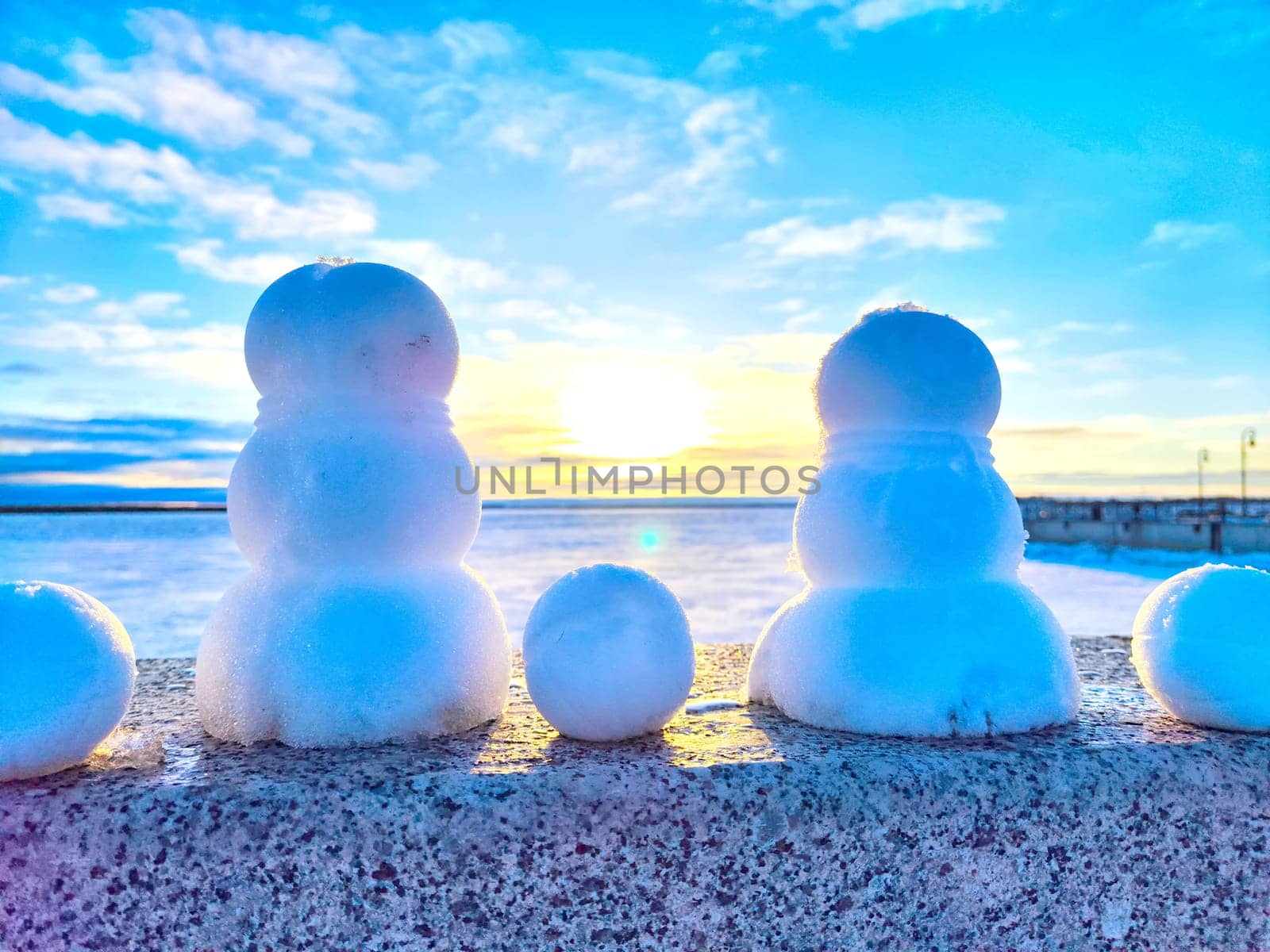 Three snowmen are positioned on a stone wall with a beautiful sunset over a frozen lake in the background.