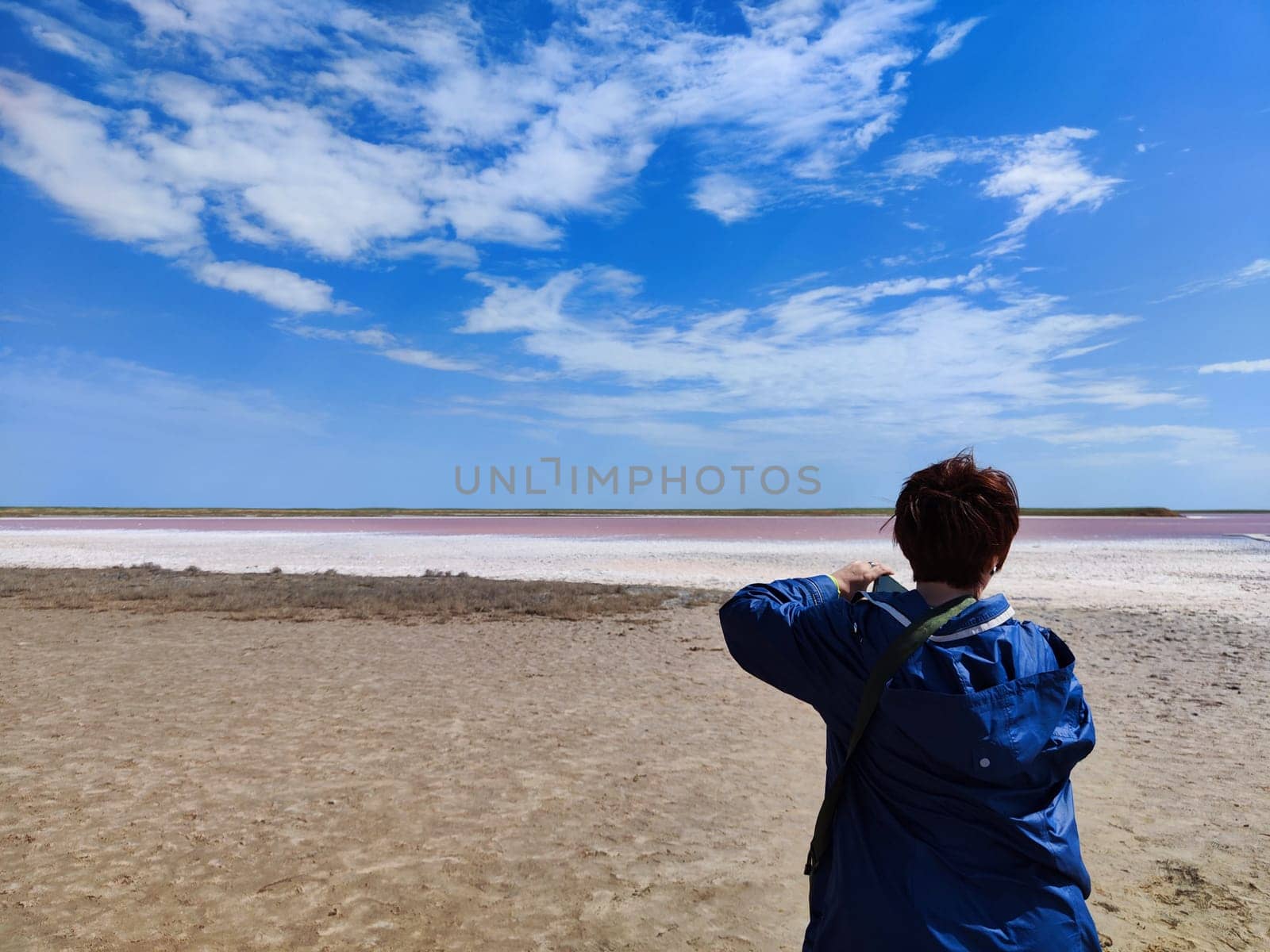 A individual stands on the sandy shore, photographing the vibrant pink salt flats beneath a bright blue sky.