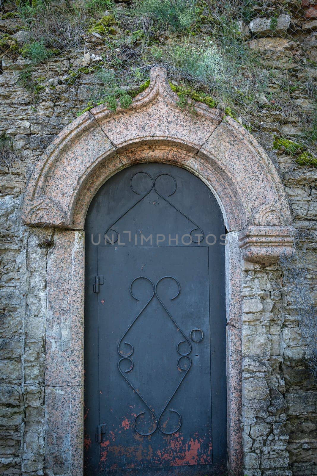 An ancient stone door with an arched opening and forged elements. Rustic architecture surrounded by moss and textured masonry creates an atmosphere of abandonment and history