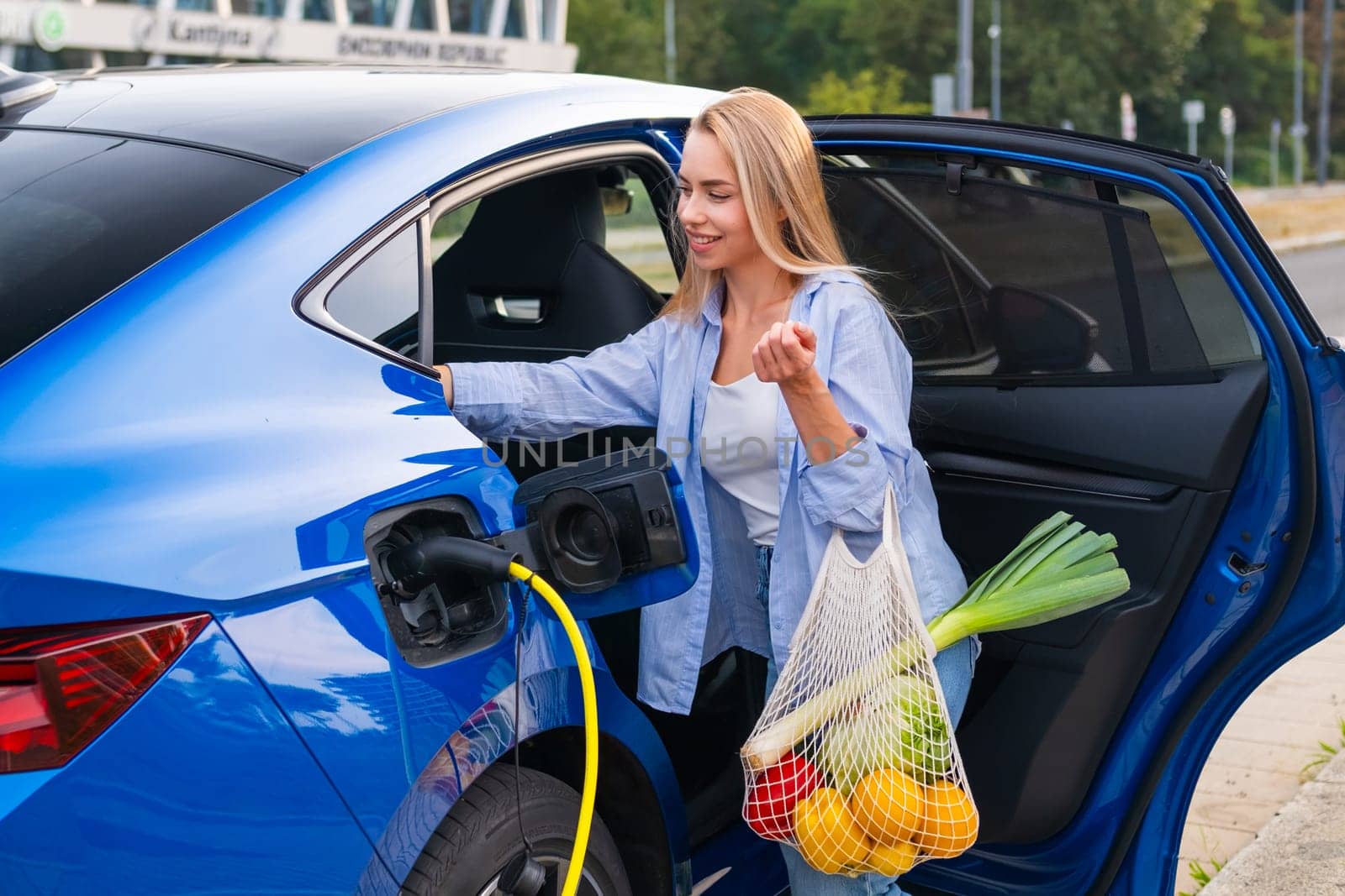 Woman is charging her electric vehicle while holding a bag of fresh groceries beside the car and places them on the back seat