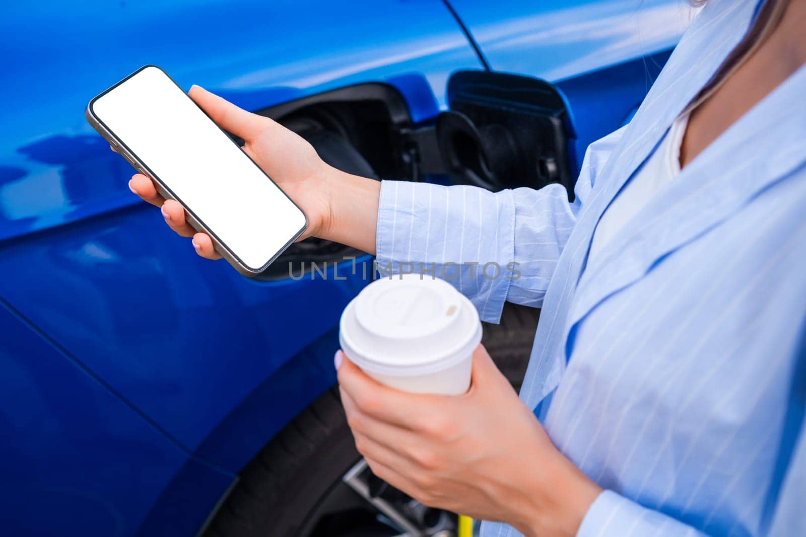 Woman charges their electric car, holding a coffee cup and checking their smartphone nearby