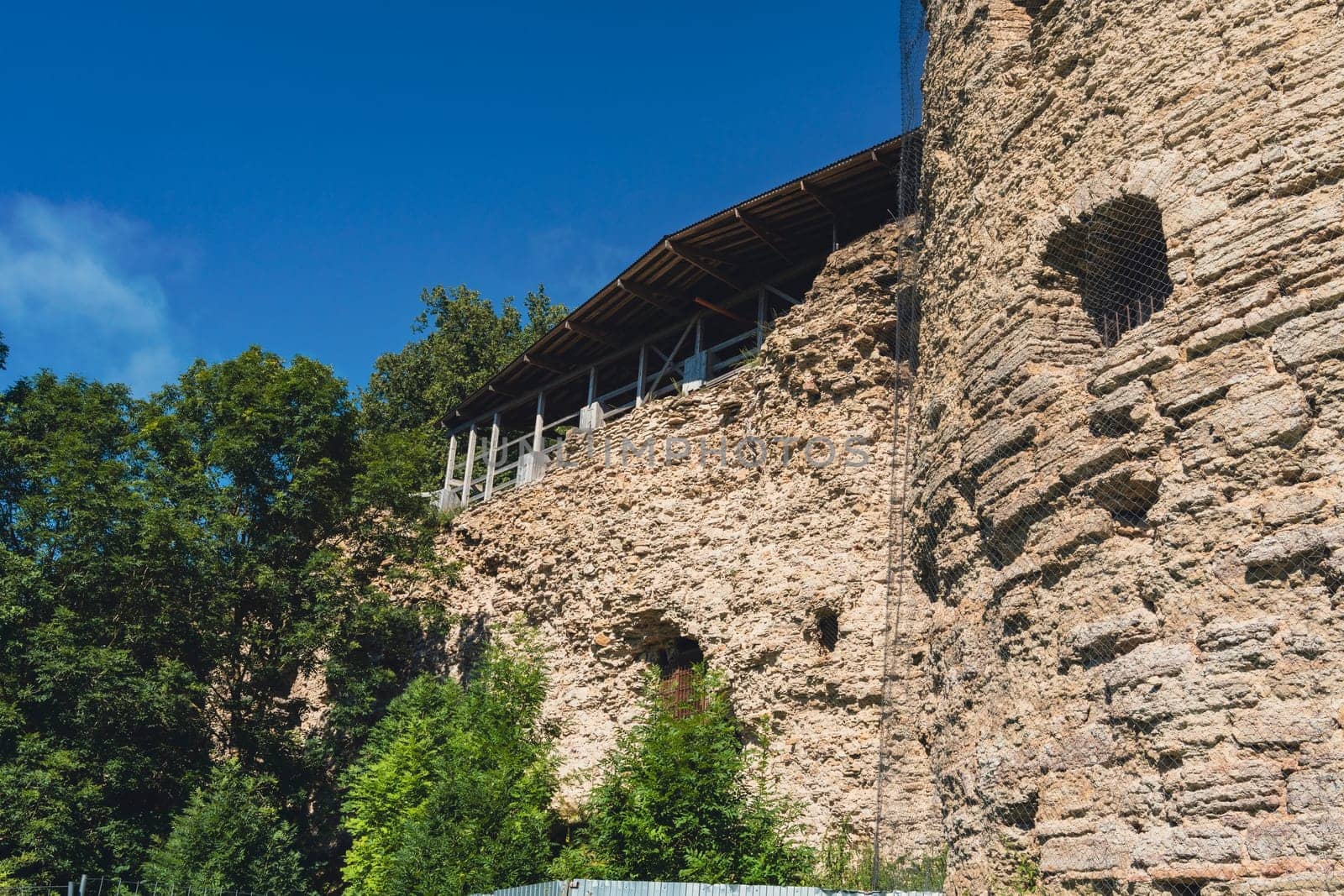 Close-up of an ancient fortress wall made of rugged stone, partially covered by greenery, with trees and a clear blue sky in the background. Historic and peaceful setting