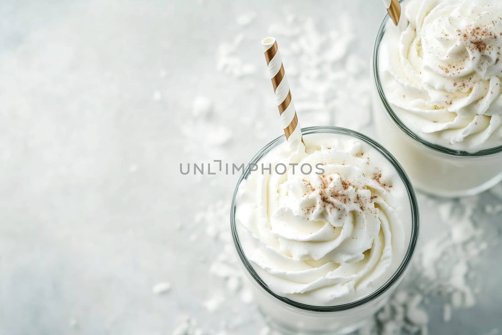 Two clear glasses filled with creamy beverages are topped with generous swirls of whipped cream, accented by striped straws. Shredded coconut decorates the surface.