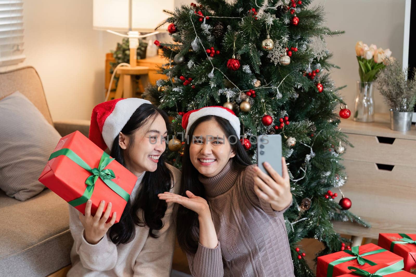 A young woman couple joyfully exchanges a surprise gift box on Christmas Day, surrounded by festive decorations in a cozy home setting.