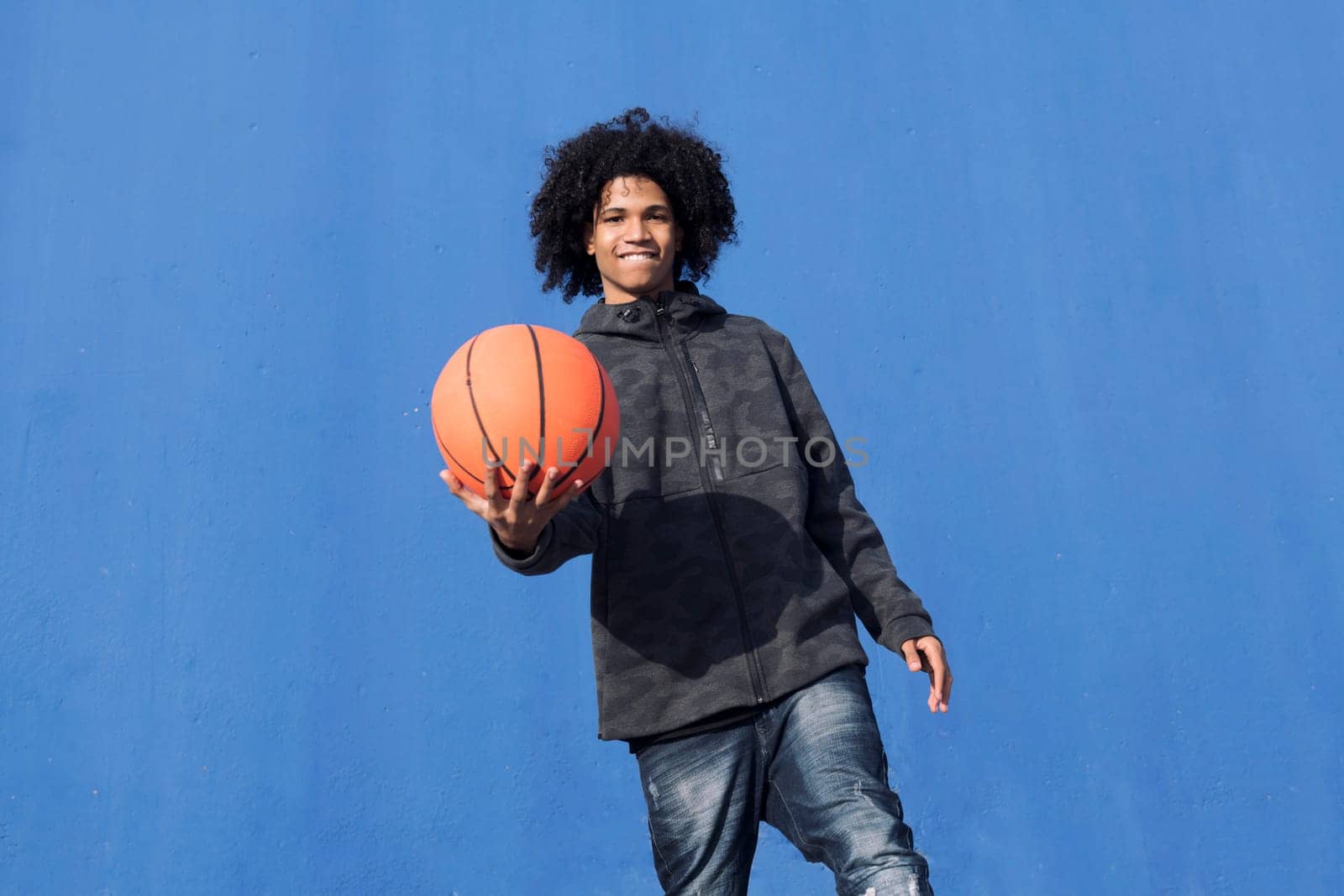positive teen african american male with curly hair offering hand with basketball ball towards camera against blue background, concept of sport and young lifestyle, copy space for text