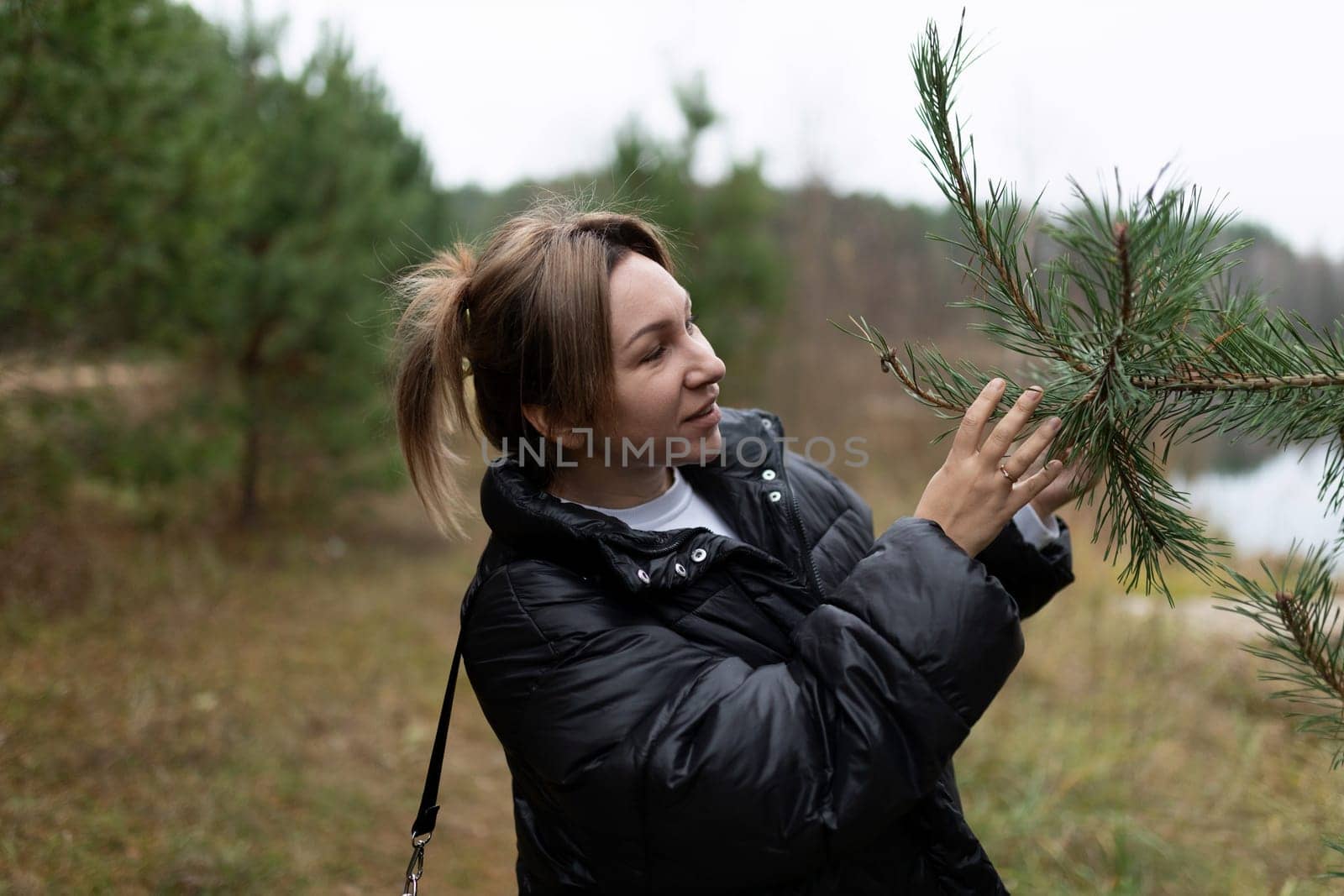 A woman in a black jacket inspects the branches of a pine tree while walking through a serene forest. The lake is visible in the background.