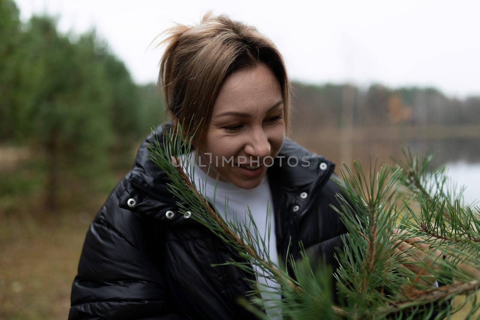 A woman is carefully collecting pine branches by the edge of a tranquil lake. The area is surrounded by lush trees under a gray, overcast sky, depicting autumn scenery.