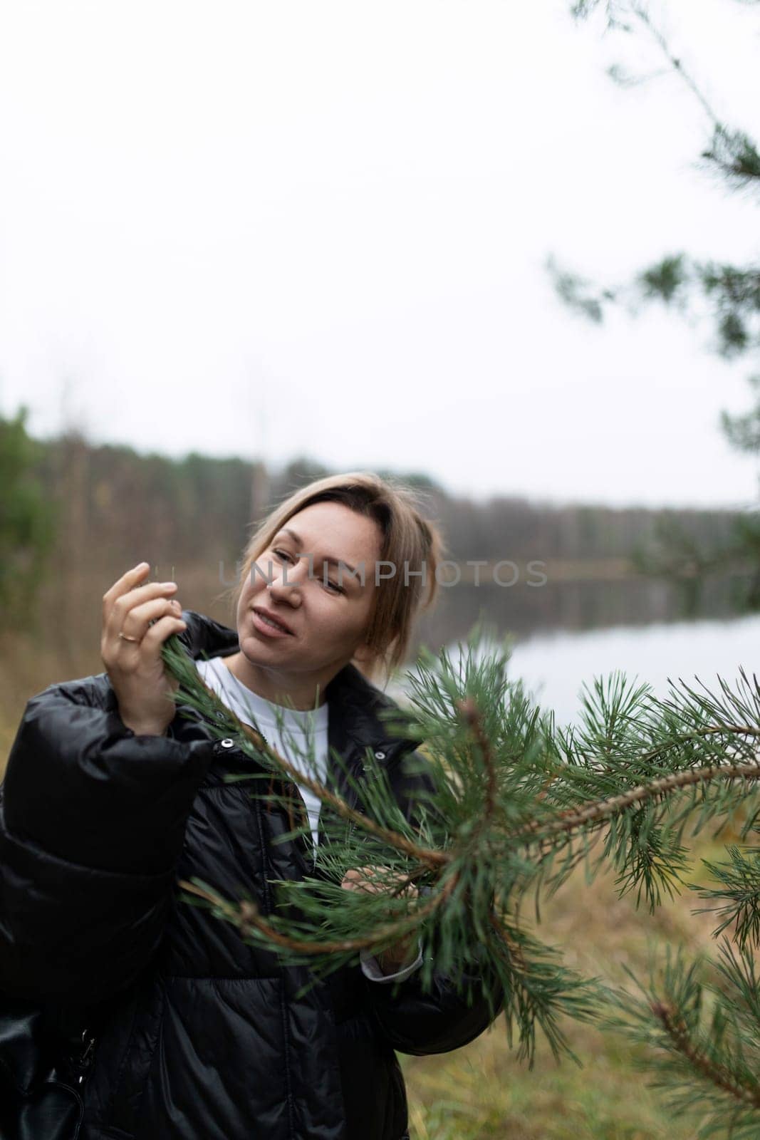 Near a tranquil lake, a woman inspects a pine branch while surrounded by lush greenery and soft autumn light, enjoying the natural environment.