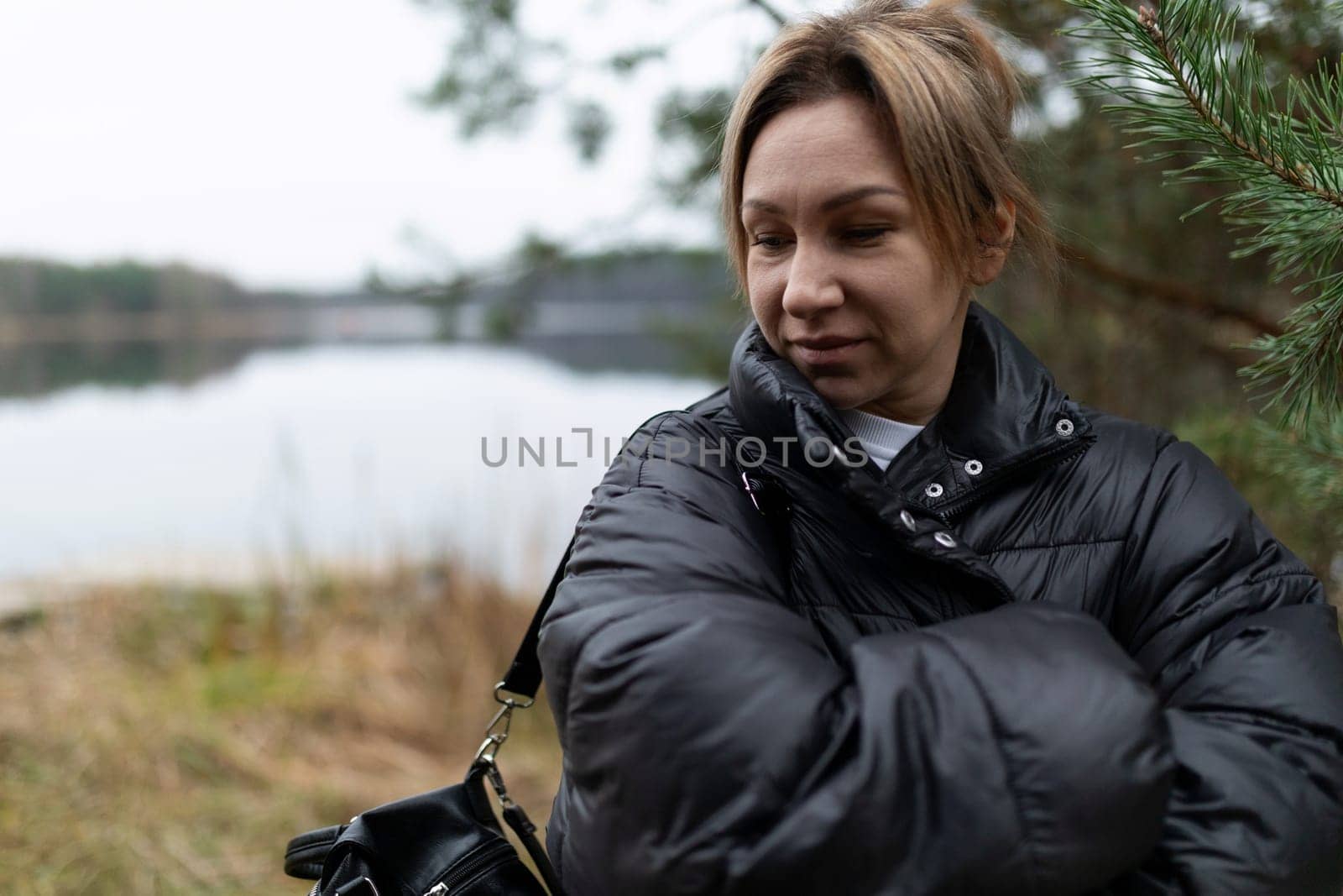 A woman with crossed arms looks contemplatively towards a serene lake, while lush greenery frames the view. The atmosphere is calm and reflective.