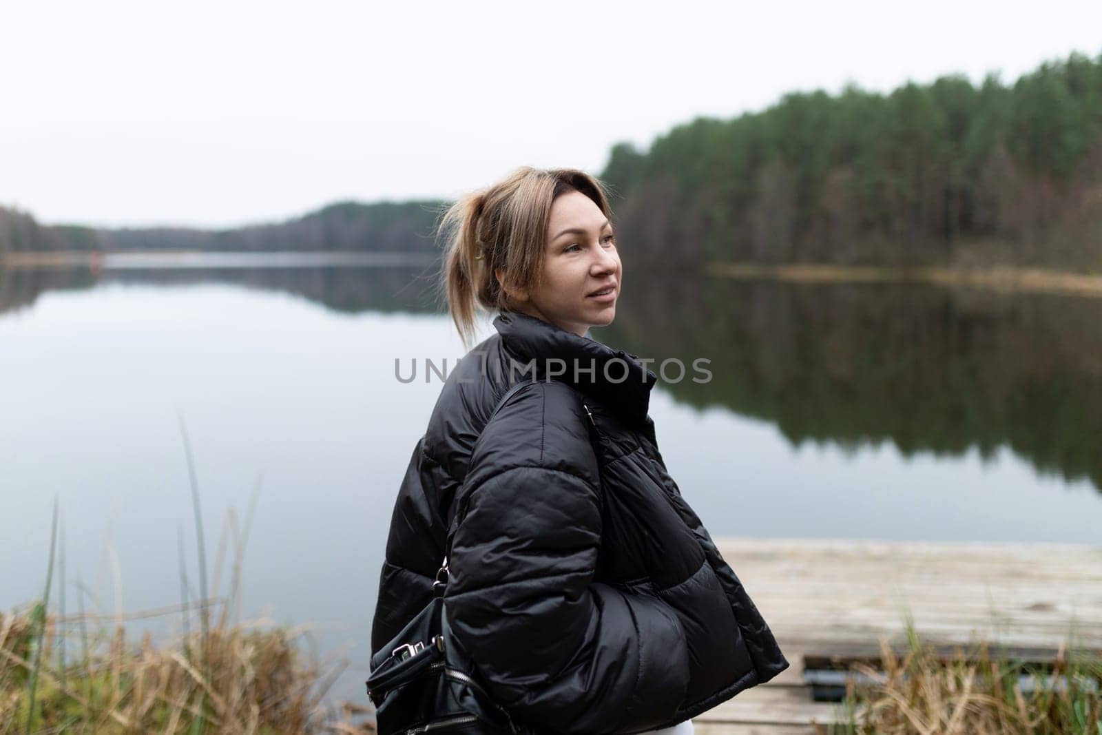 A young woman stands peacefully by a calm lake, wearing a warm jacket as she gazes thoughtfully at the serene surroundings of green trees and still water.