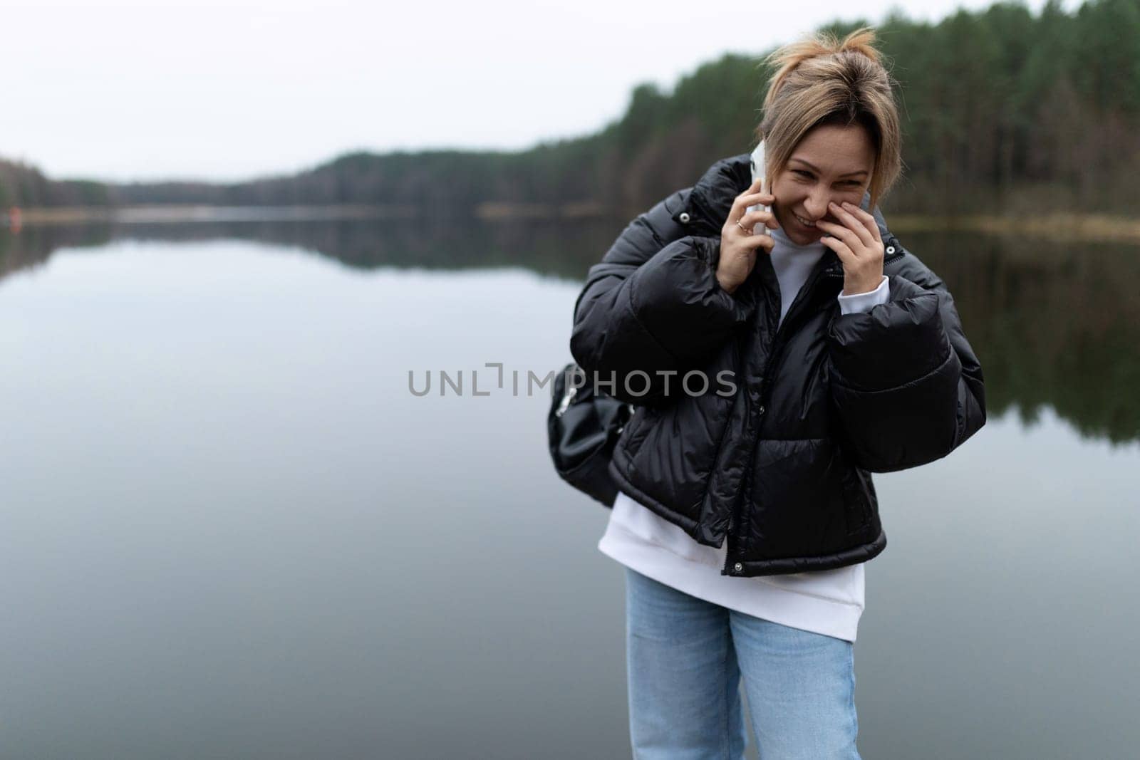 A young woman wearing a black puffer jacket and jeans laughs while speaking on a mobile phone. She stands near a large body of water, with trees on the opposite bank of the lake.