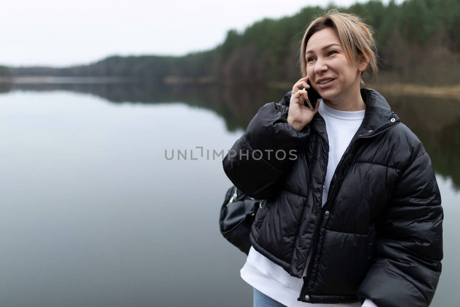 A young woman wearing a black puffer jacket and white shirt stands near a lake on a cloudy day. She is talking on her phone with a smile on her face. Her backpack is slung over her shoulder.