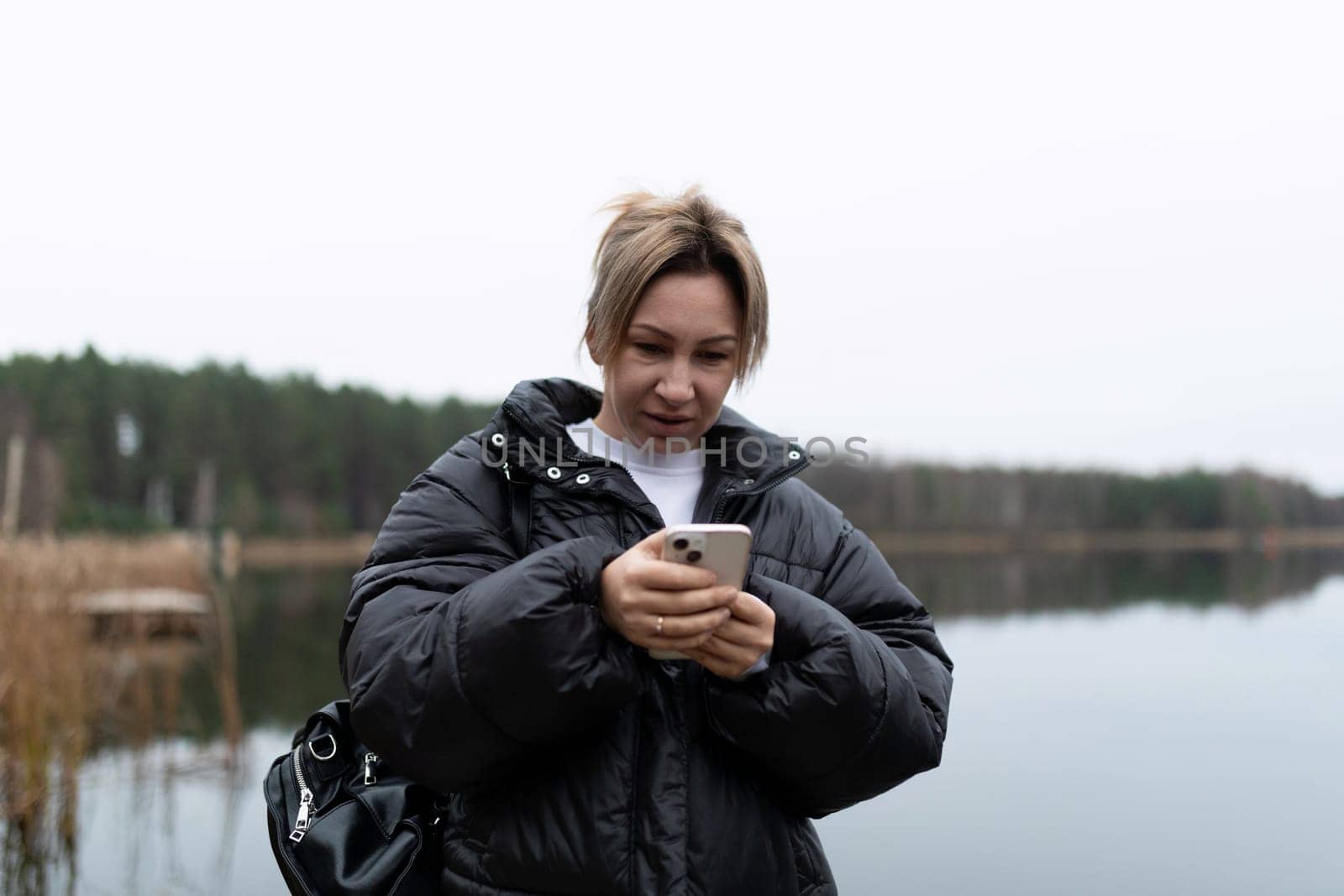 A woman with short blonde hair is standing by a lake. She is wearing a black puffer jacket and is looking at her phone. The weather is cloudy, and the lake is calm.