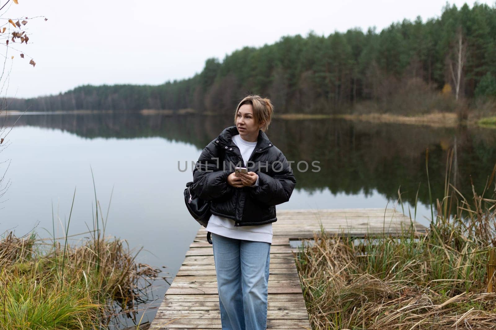 A woman in a black jacket and blue jeans walks along a wooden dock over a calm, reflective lake. She holds a smartphone in her hands and is looking towards the right side of the frame.