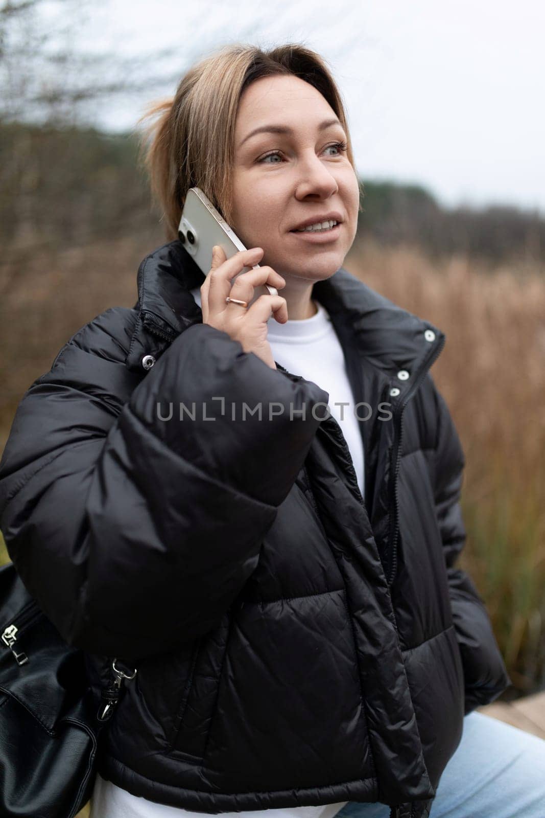 A woman with blonde hair, wearing a black puffer jacket and a white shirt underneath, holds a phone to her ear while talking.