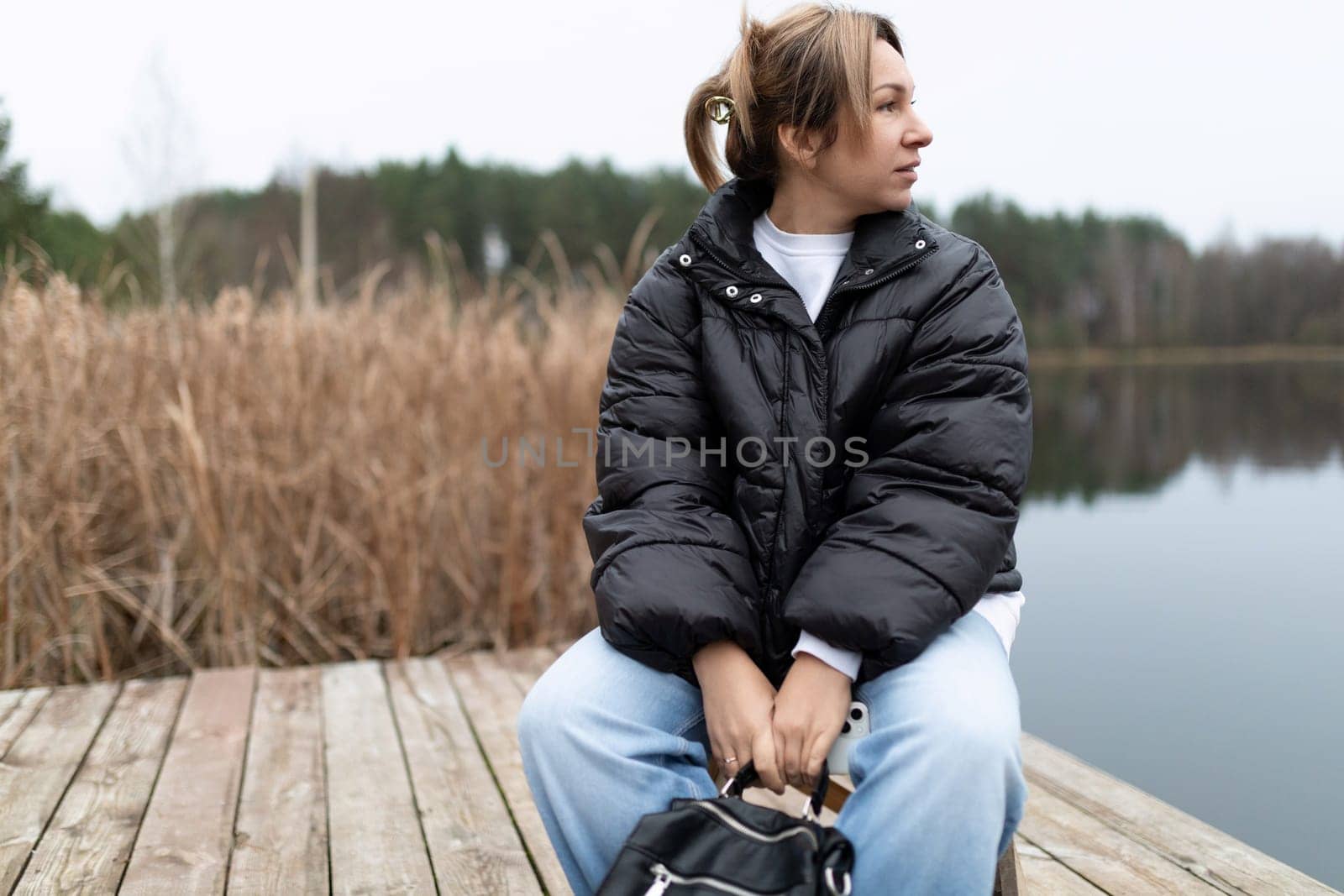 A woman wearing a black puffer jacket and blue jeans sits on a wooden dock. She is looking out at a lake, her hands resting on a black bag in her lap. There is a field of tall grass behind her.