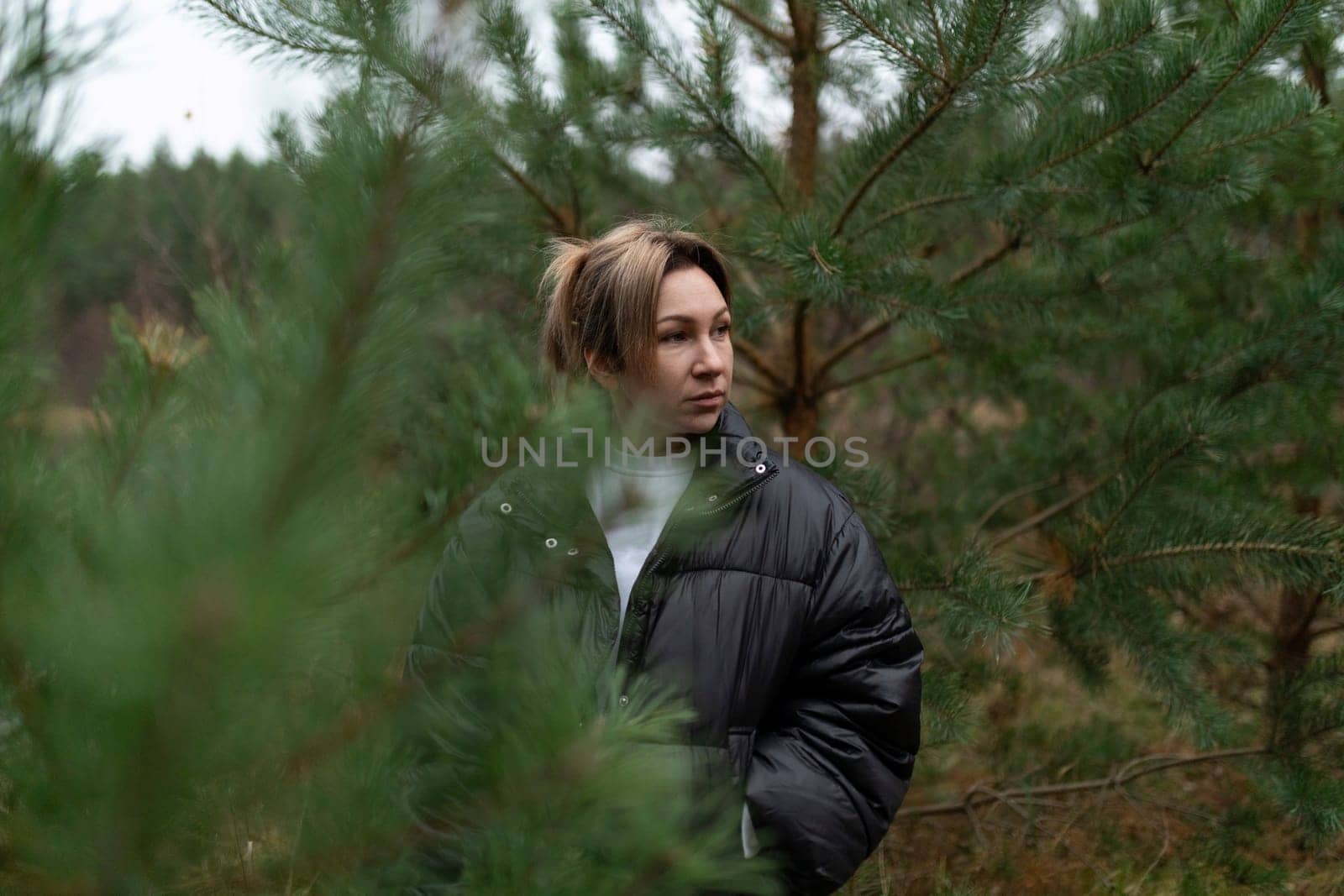 A woman in a black jacket observes her surroundings in a serene forest setting, surrounded by lush green trees, capturing a moment of peace and introspection.