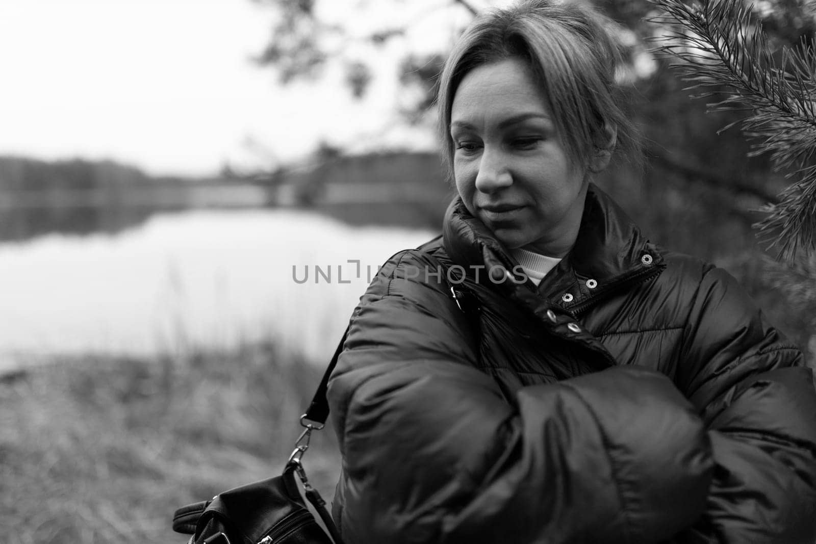 A woman wearing a warm puffy jacket crosses her arms while contemplating by a serene lake surrounded by gentle nature. The day feels peaceful and reflective.
