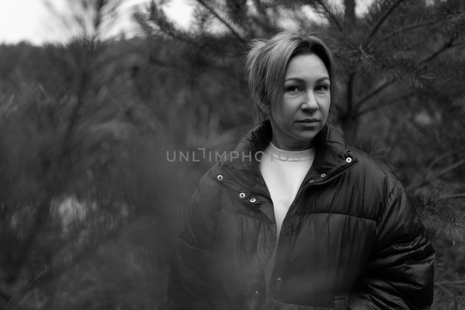 In a tranquil forest, a woman wearing a black jacket stands quietly between evergreen trees, gazing thoughtfully into the distance on a cloudy day.