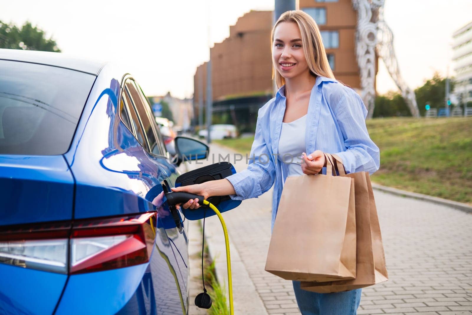 Portrait of blonde woman plugs in her electric vehicle to charge while holding multiple shopping bags.