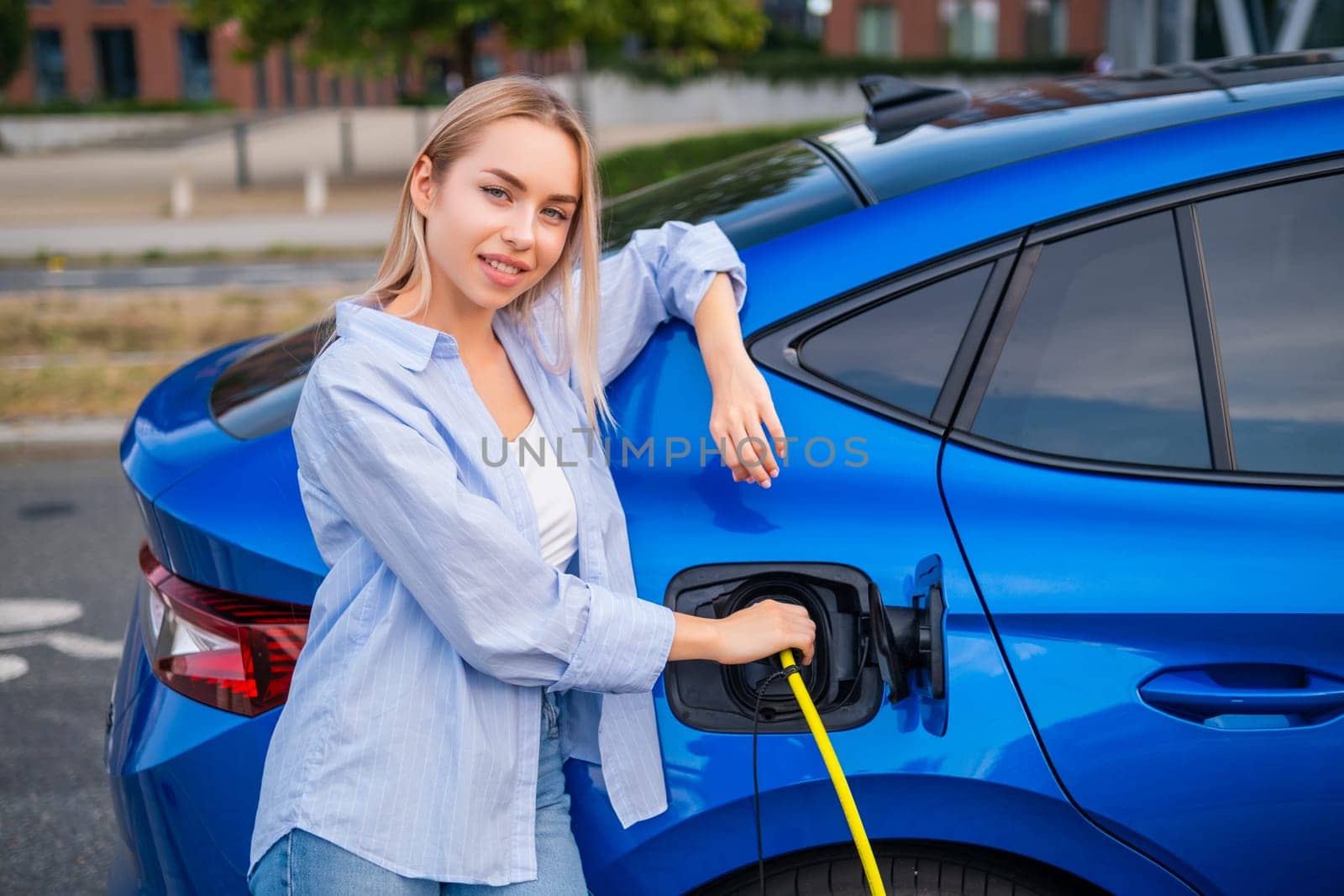 Portrait of blonde woman in casual clothes charging her electric car at a public parking station