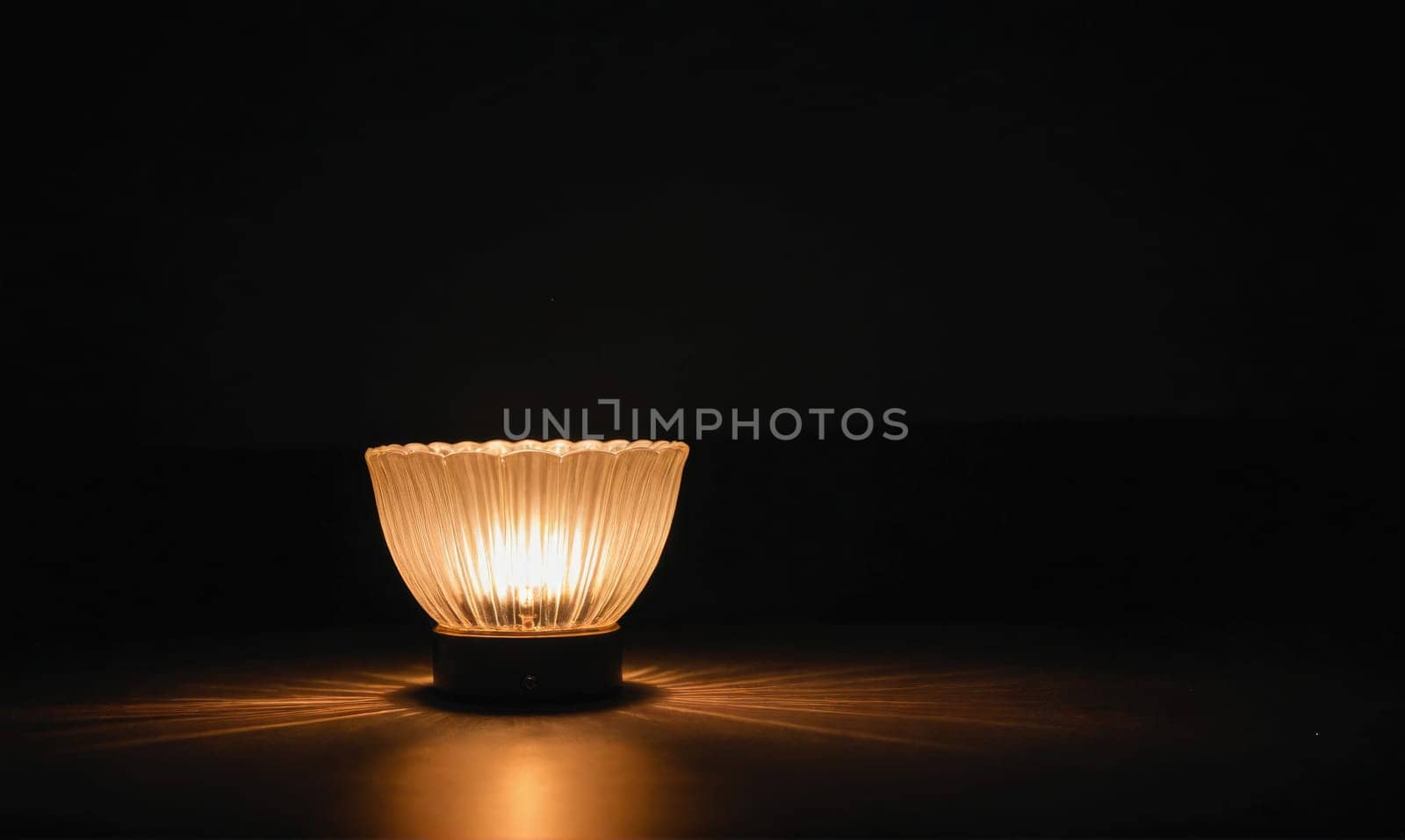 A small lamp with a glass shade illuminates a dark room, casting a warm, golden light on the table surface.