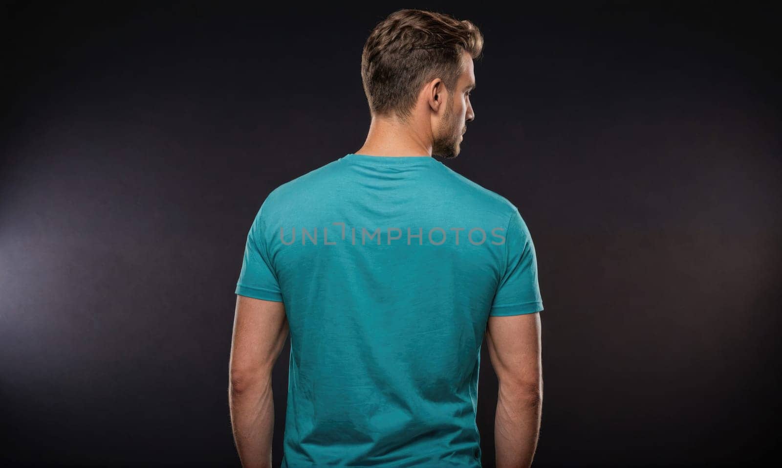 A man is standing in front of a dark background, looking over his shoulder. He is wearing a plain, light blue t-shirt. His arms are at his sides, and his hands are in his pockets.